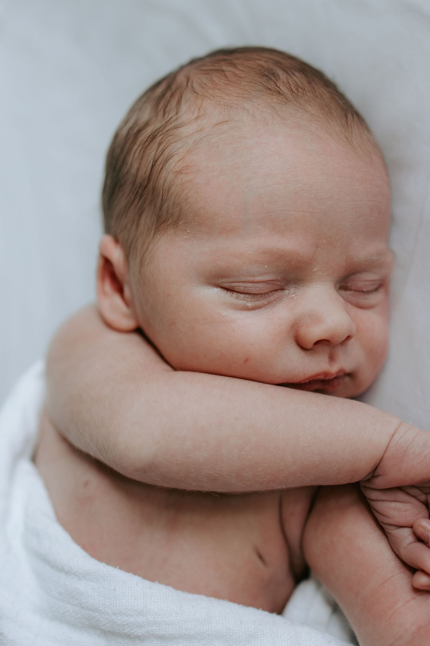 A peaceful sleeping newborn baby wrapped in a white blanket with tiny hands tucked under its chin.