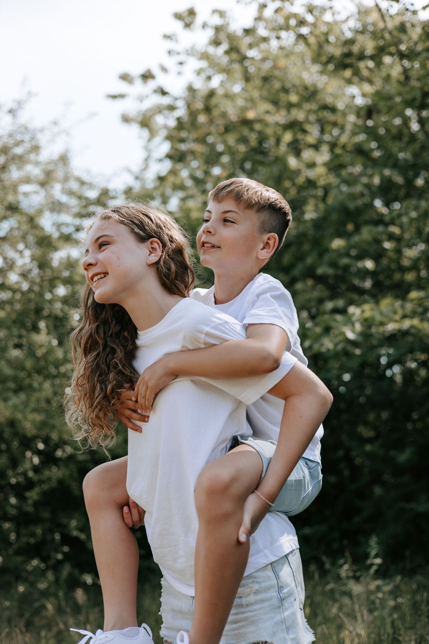 Smiling teenage girl carrying boy on her back in sunny green outdoor setting.