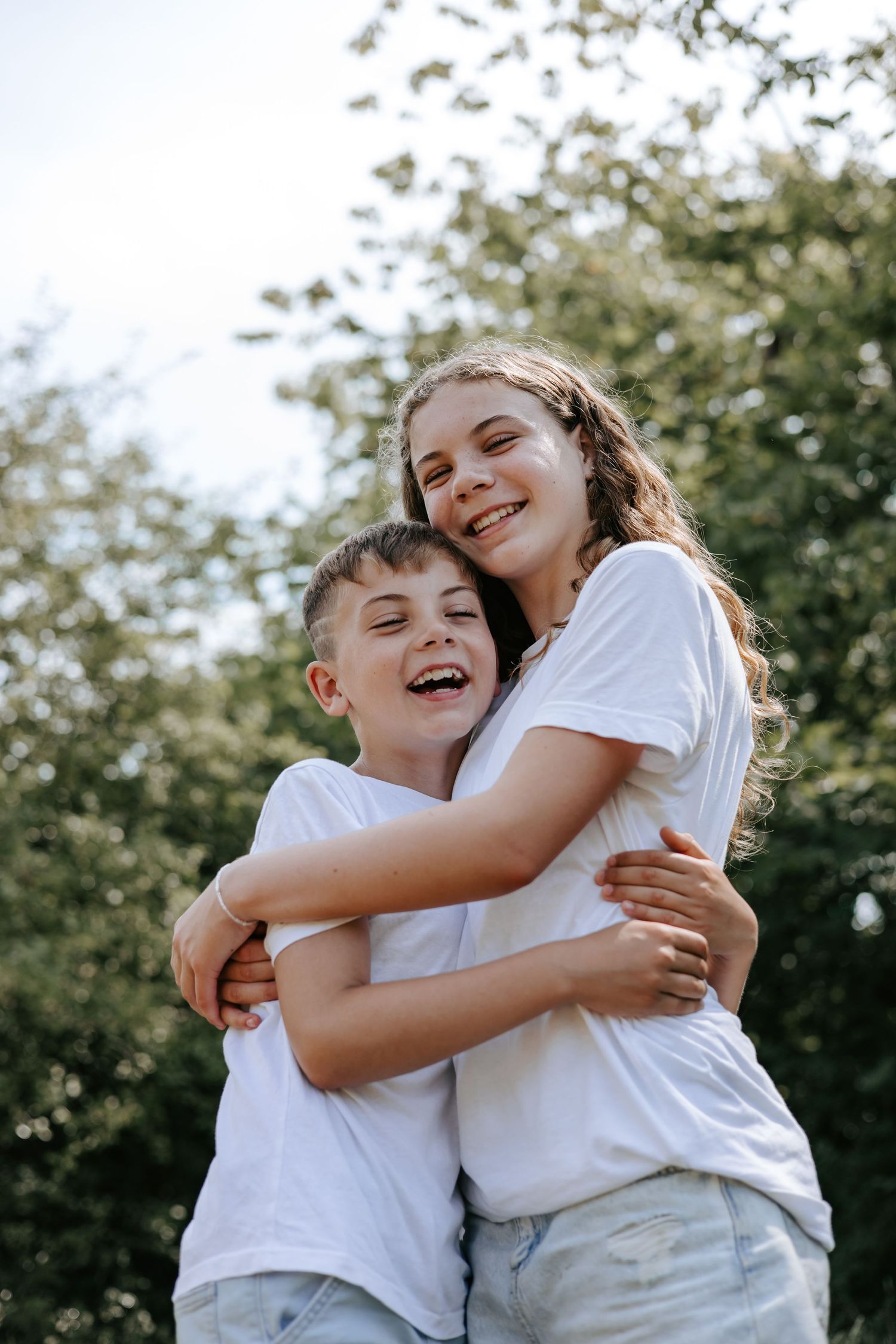 Two siblings in white t-shirts hugging and laughing together outdoors in a sunny park.