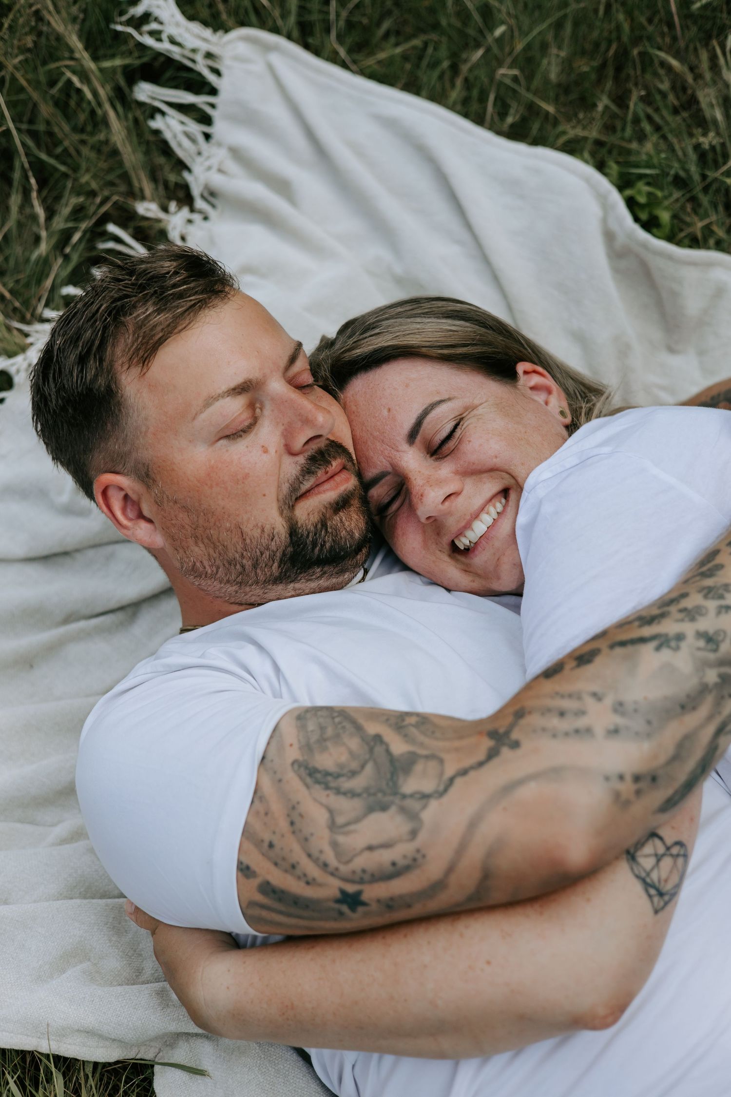 Affectionate tattooed couple cuddling and smiling while lying on blanket in grass.
