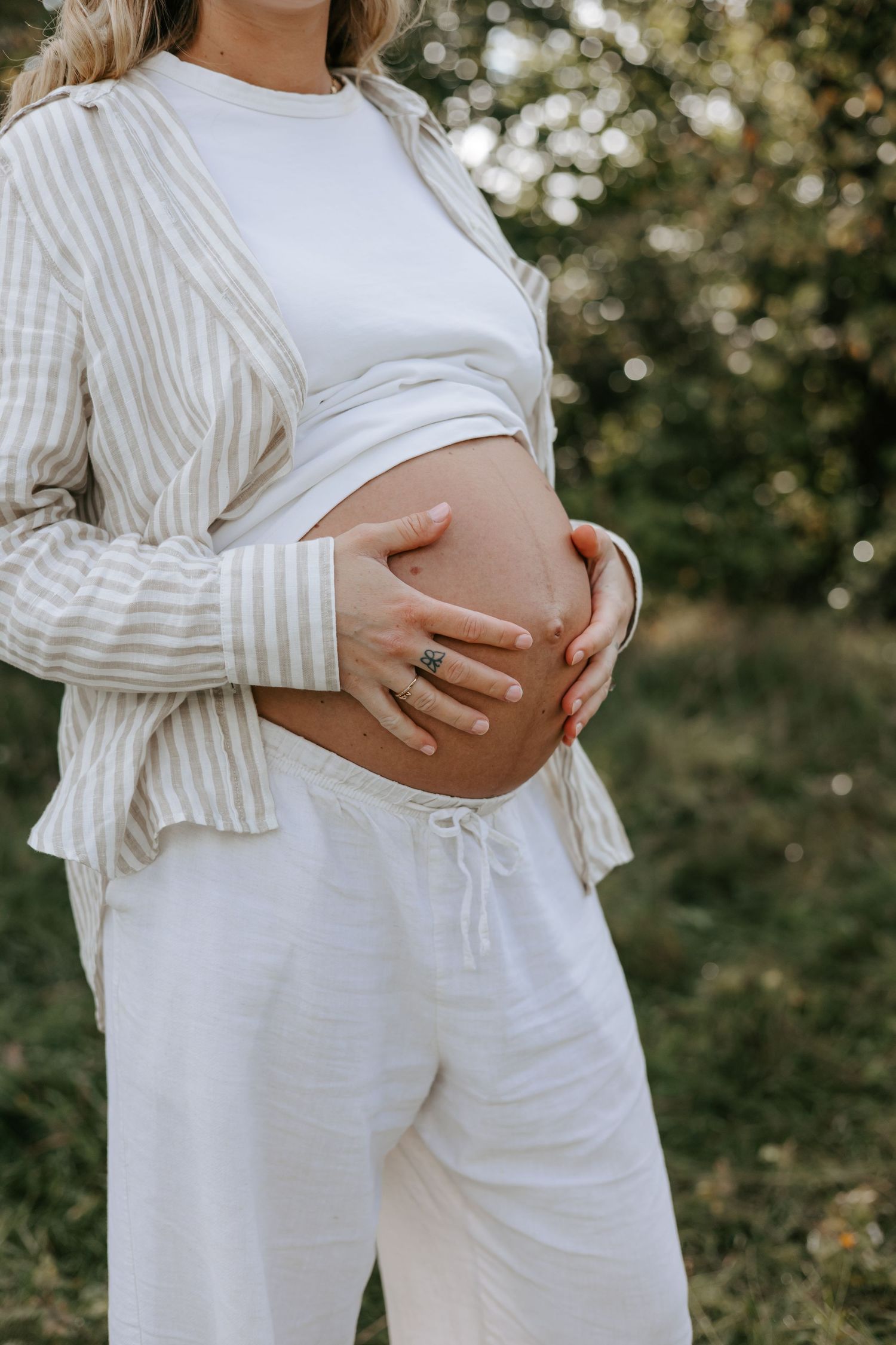 Pregnant woman in white pants and striped shirt cradling her baby bump outdoors in a natural green setting.