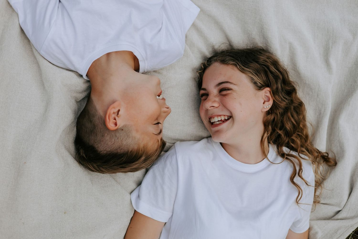 Two cheerful children lying on a blanket laughing together in a bright outdoor setting.