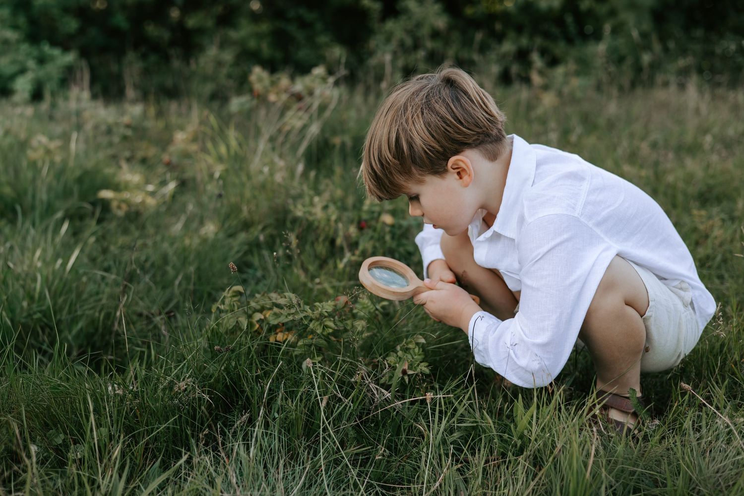 Young boy crouching in green grass, looking through a magnifying glass at plants outdoors.