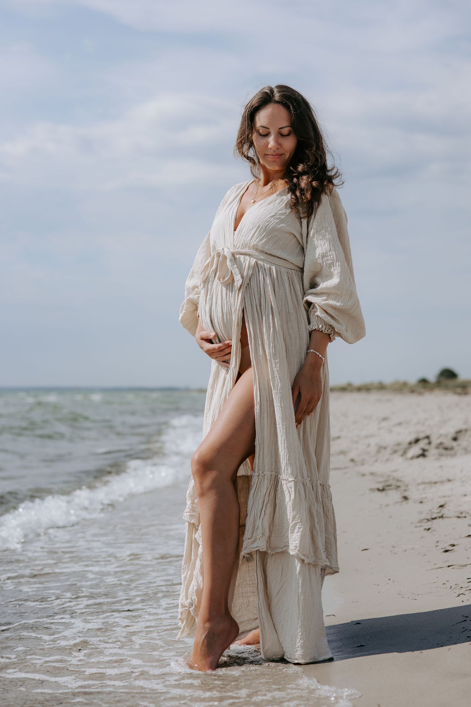 Pregnant woman in elegant beige maxi dress cradling belly on sandy beach shore.