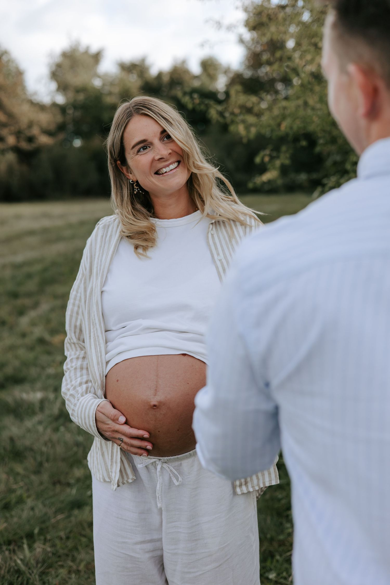 Smiling pregnant woman in white outfit holds her baby bump while standing outdoors with a man in a park.