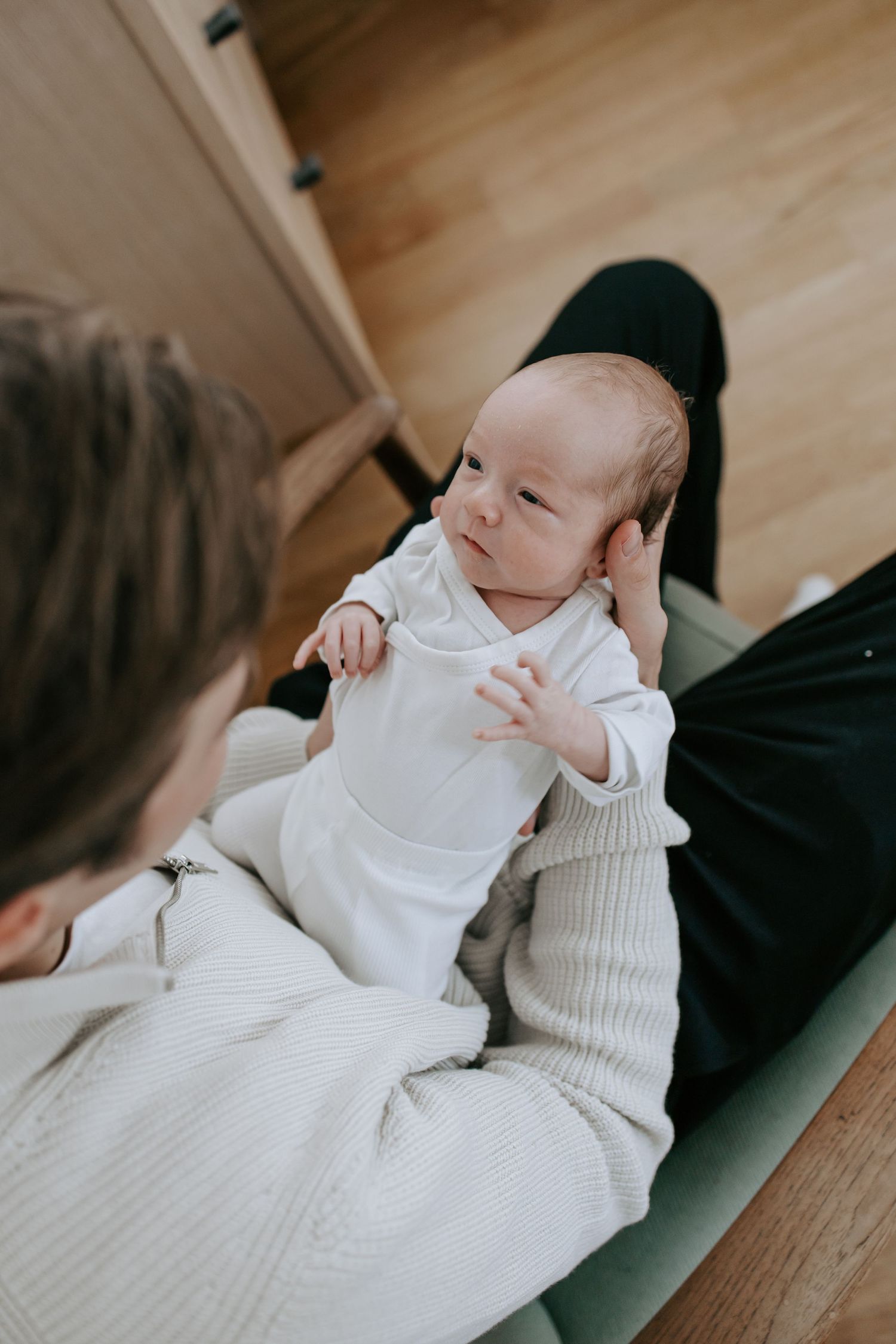 Newborn baby in white onesie lying on mother's lap looking up alertly.