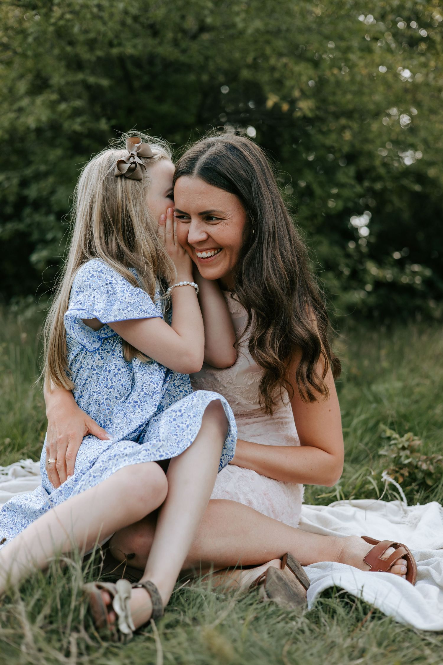 Young blonde girl whispering to her smiling mother while sitting on a blanket in grass.