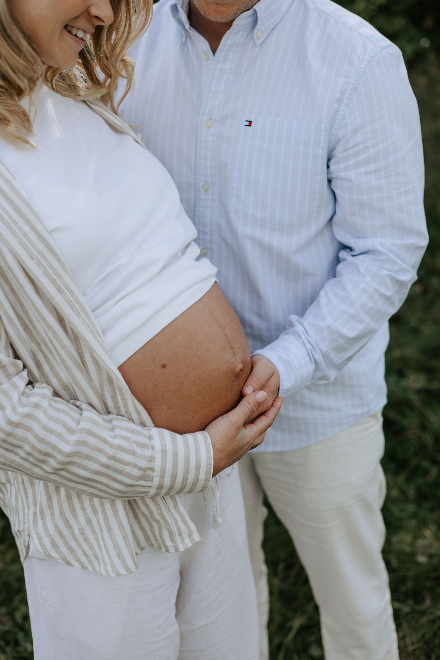 Couple sharing a tender moment outdoors, with man in Tommy Hilfiger shirt gently touching pregnant woman's bare belly.