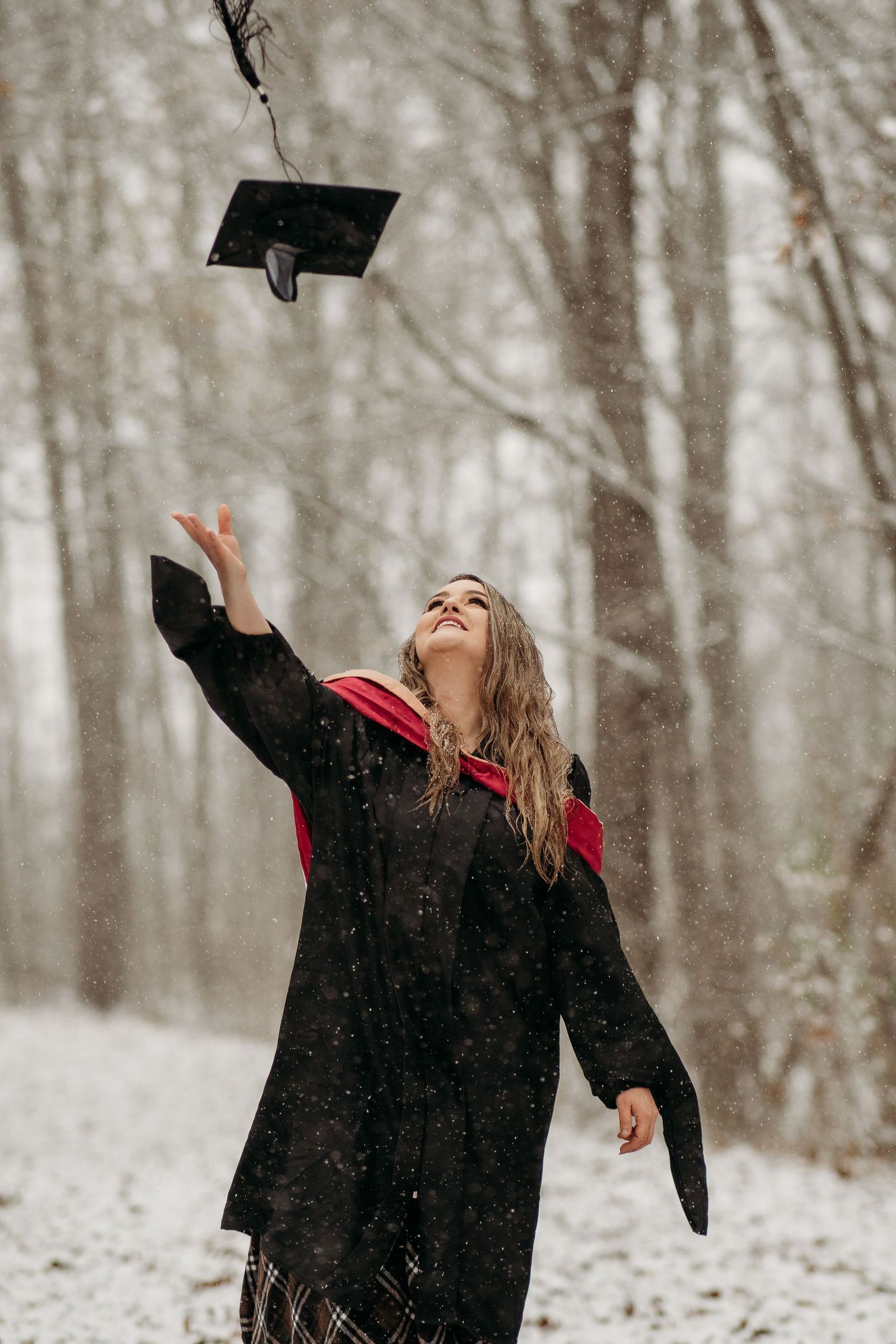 Graduate in black cap and gown tossing graduation cap in snowy winter forest setting.