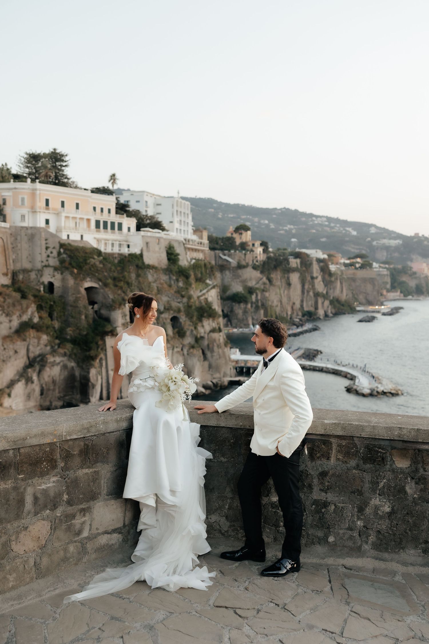 Bride and groom hold hands on a scenic Sorrento cliffside terrace overlooking the sea on their wedding day.