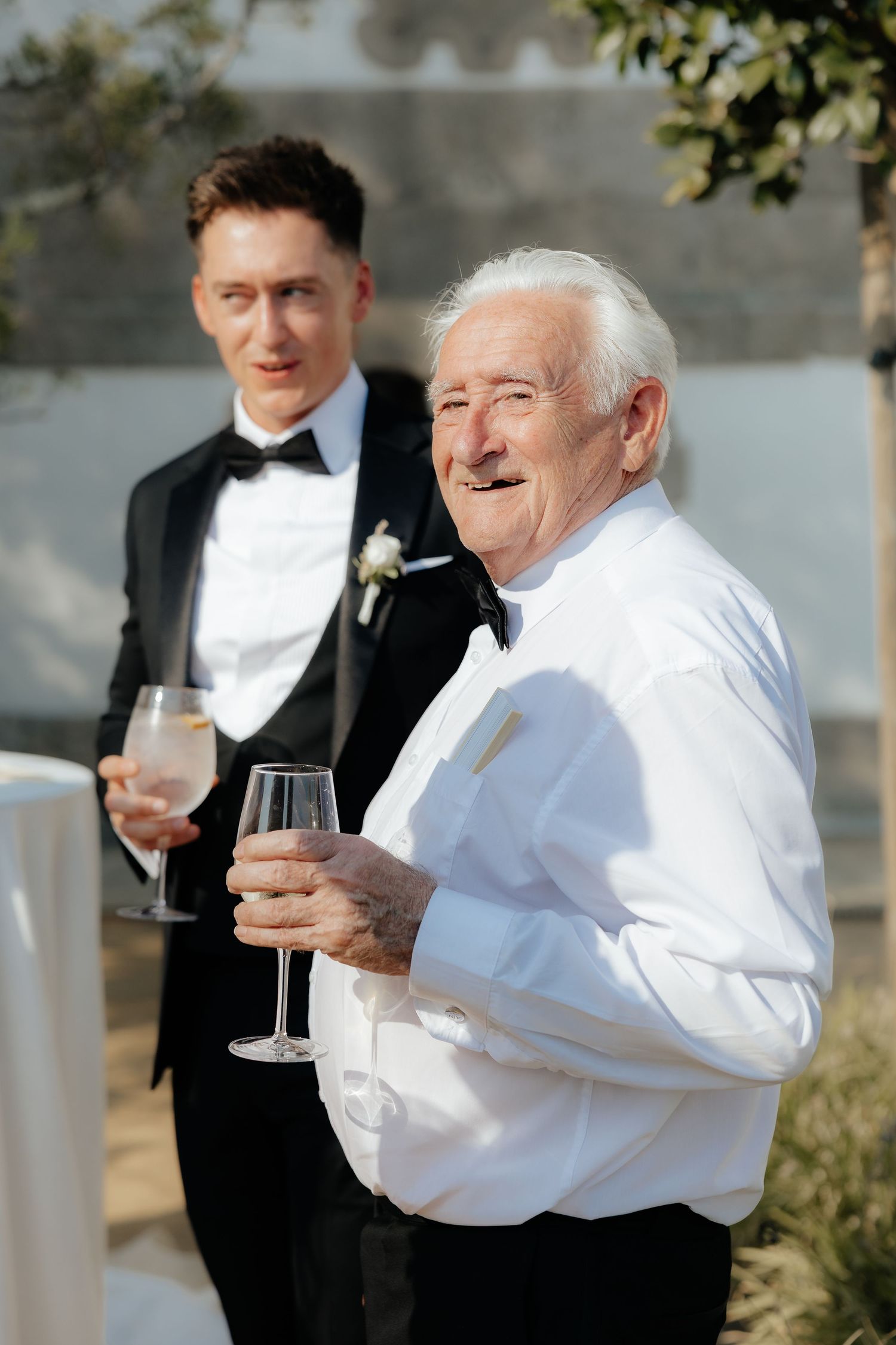 Two guests at a formal event share a moment while holding wine glasses, one in a tuxedo and one in a white dress shirt.
