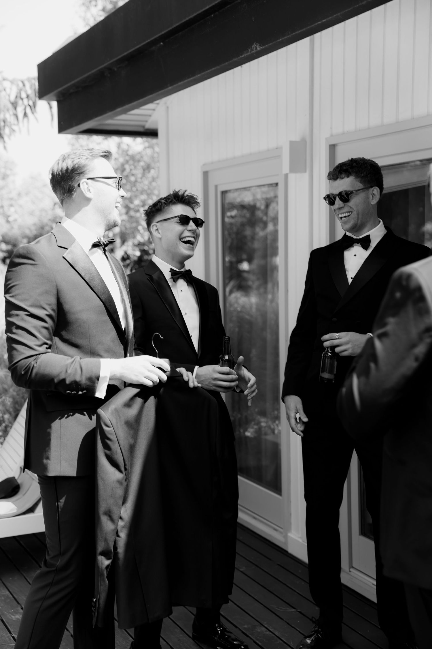 Three groomsmen in formal suits and sunglasses share a laugh together on a deck in black and white photography.