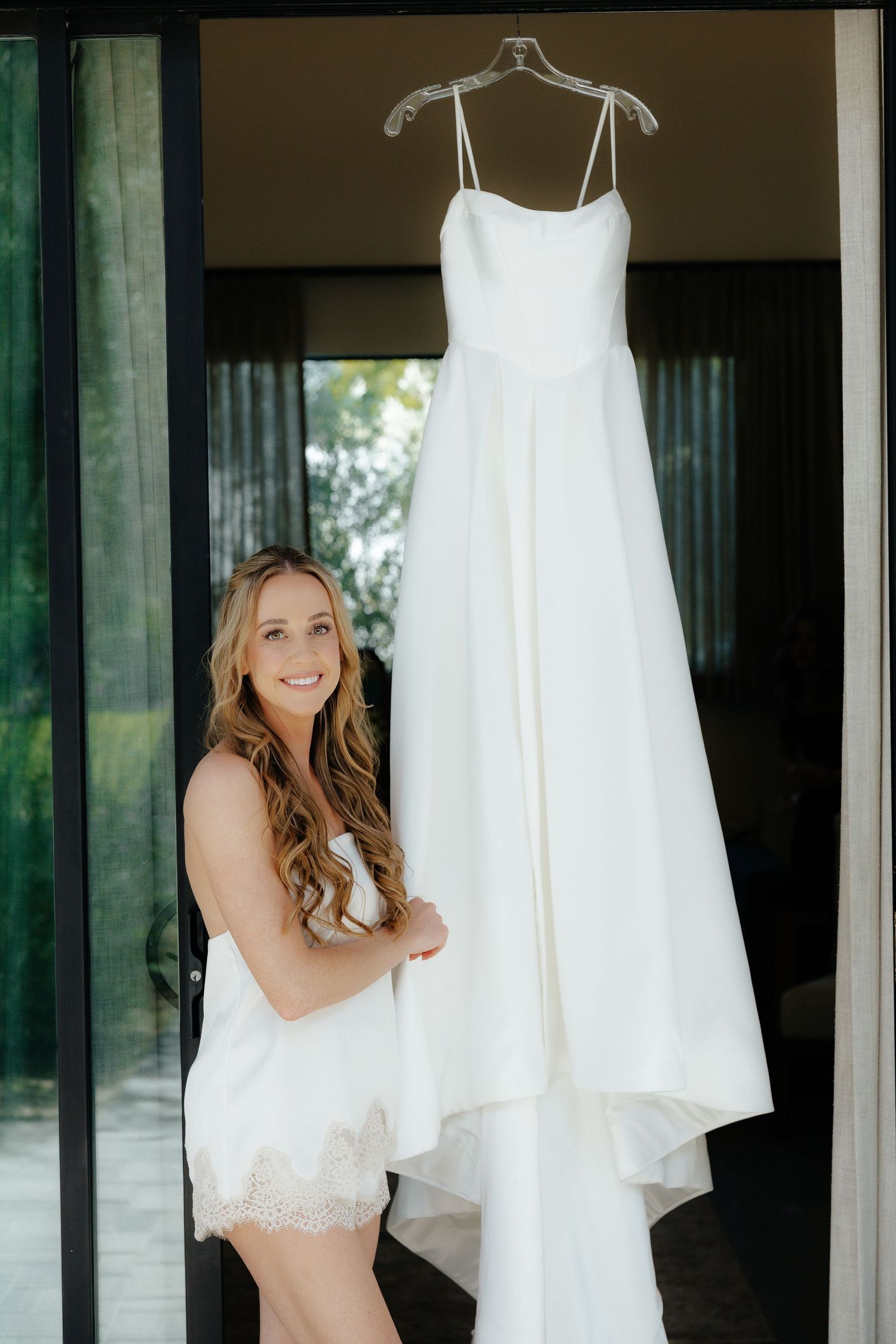 Smiling bride in white lace romper standing beside her hanging wedding gown in doorway.