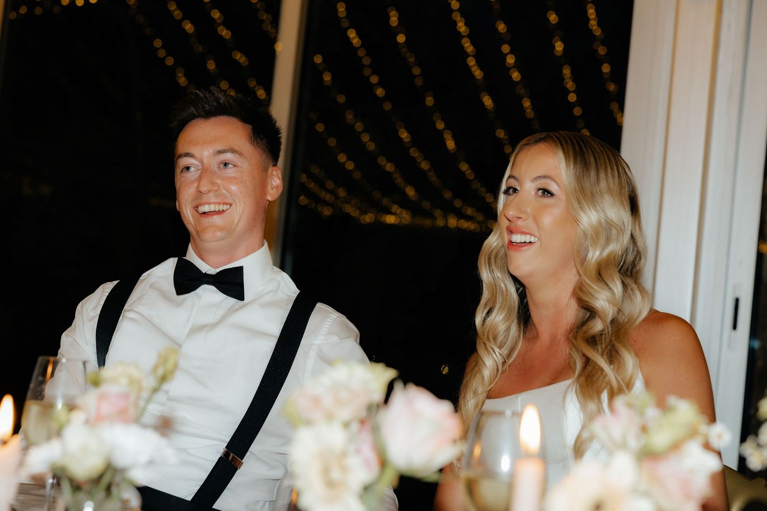 A couple shares a candid laughing moment at an elegant evening reception with twinkling lights in the background.