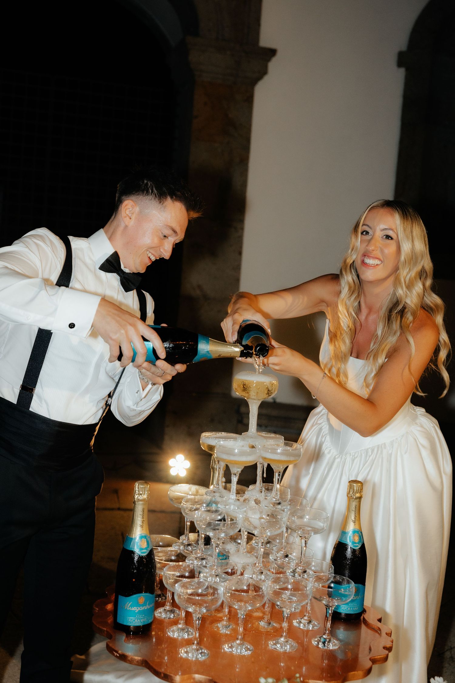 Couple pouring champagne into a tower of stacked glasses during a wedding celebration.