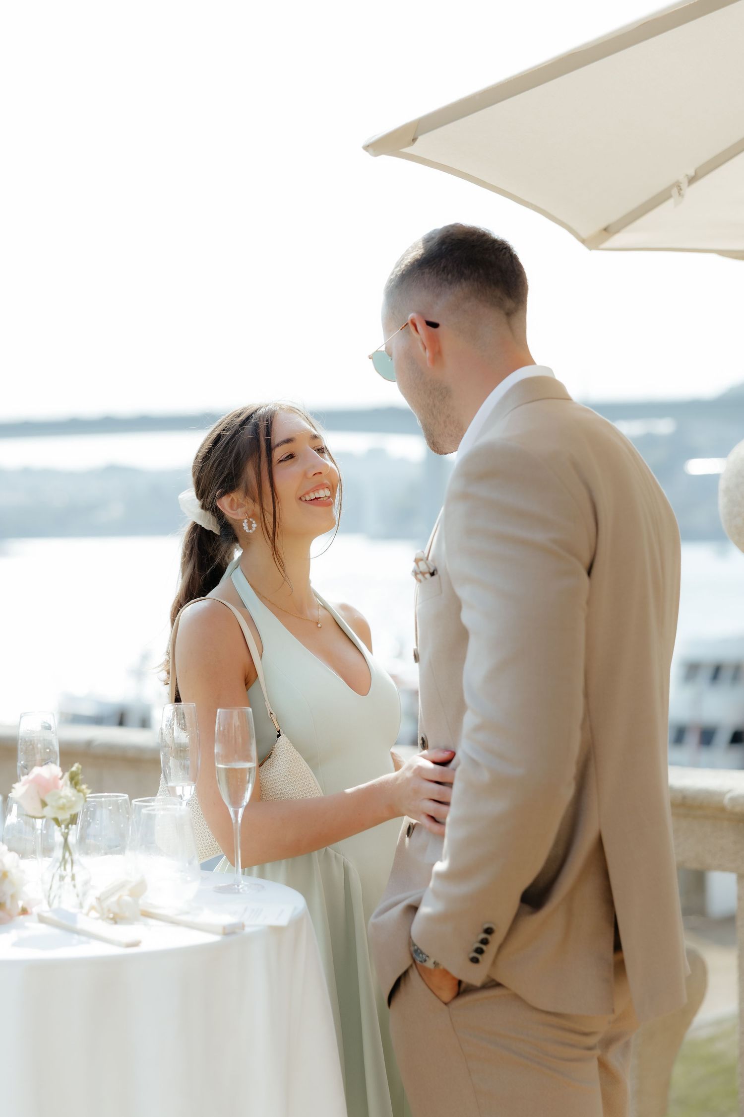 A couple shares a romantic moment at a waterfront wedding celebration with champagne glasses and floral decor.