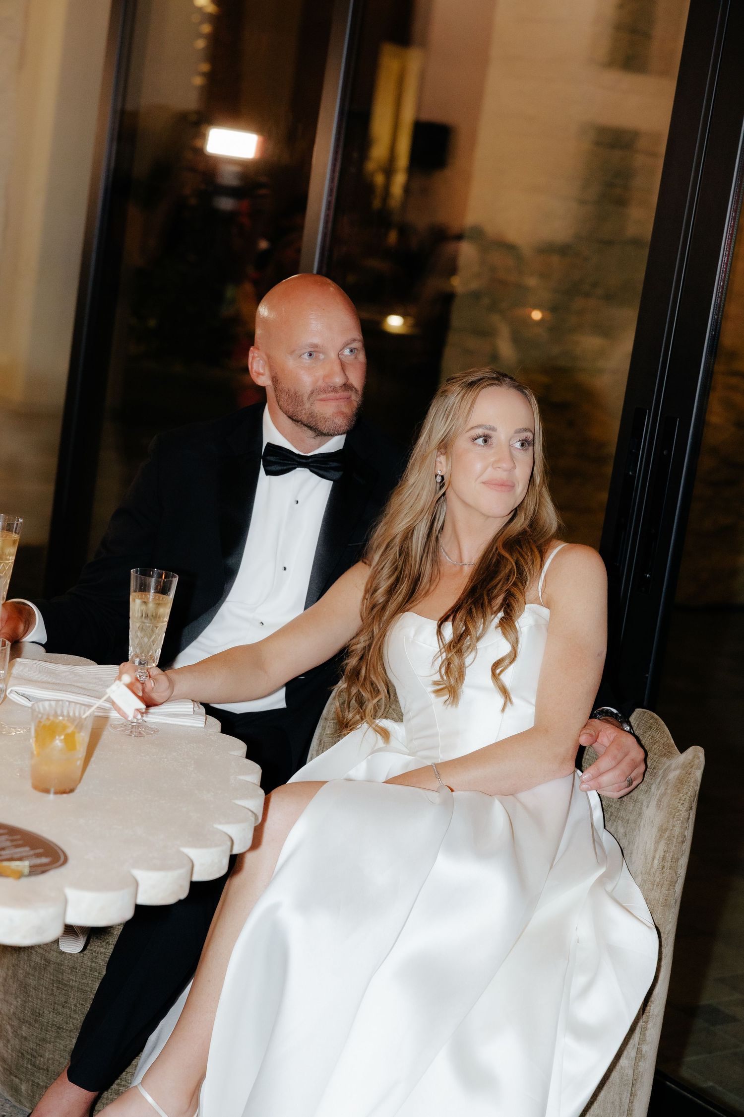 Bride and groom sit together at their wedding reception table, dressed in white gown and black tuxedo with bow tie.