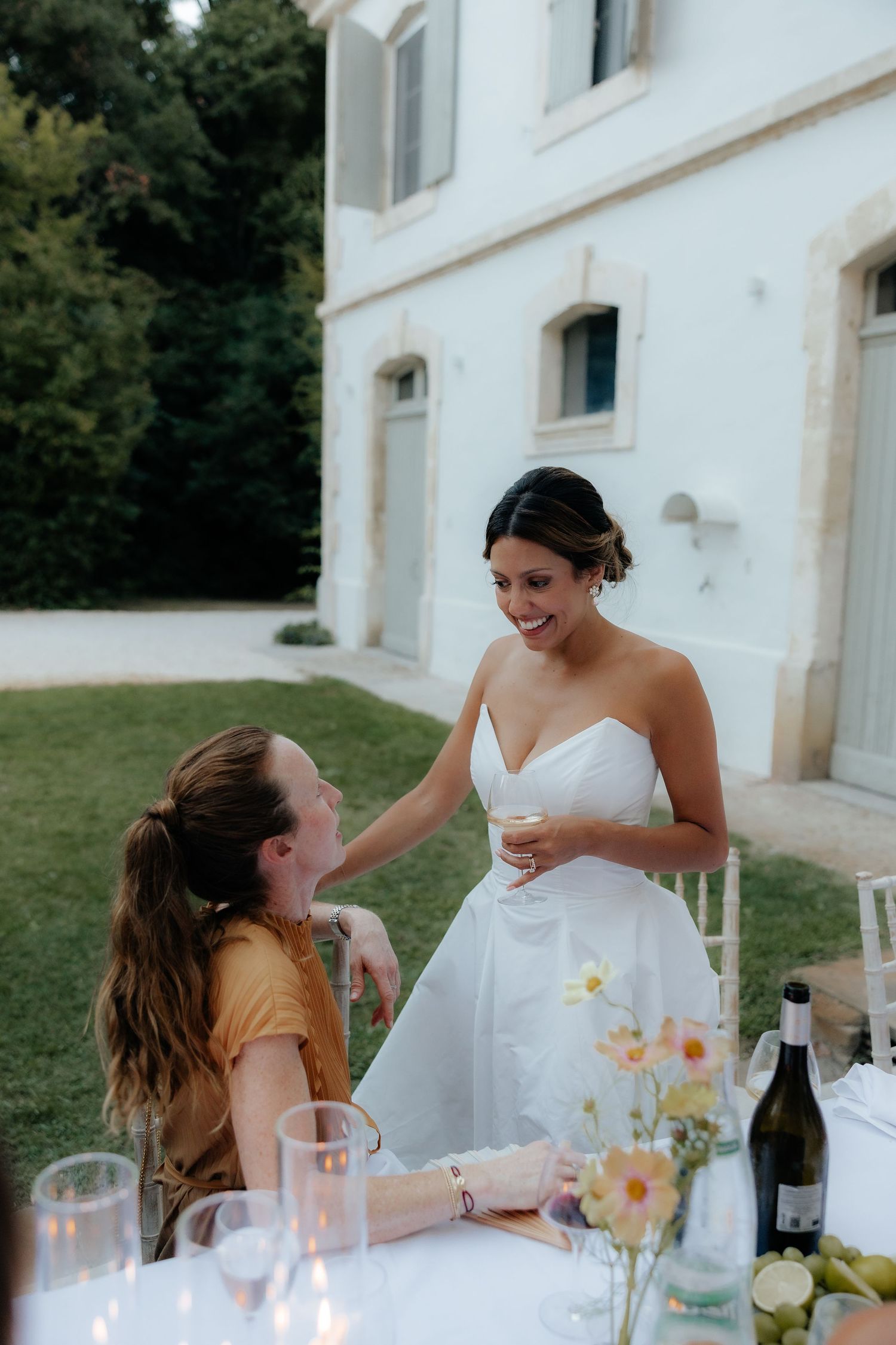 A joyful moment shared between friends at an outdoor wedding reception with floral centerpieces and wine bottles.