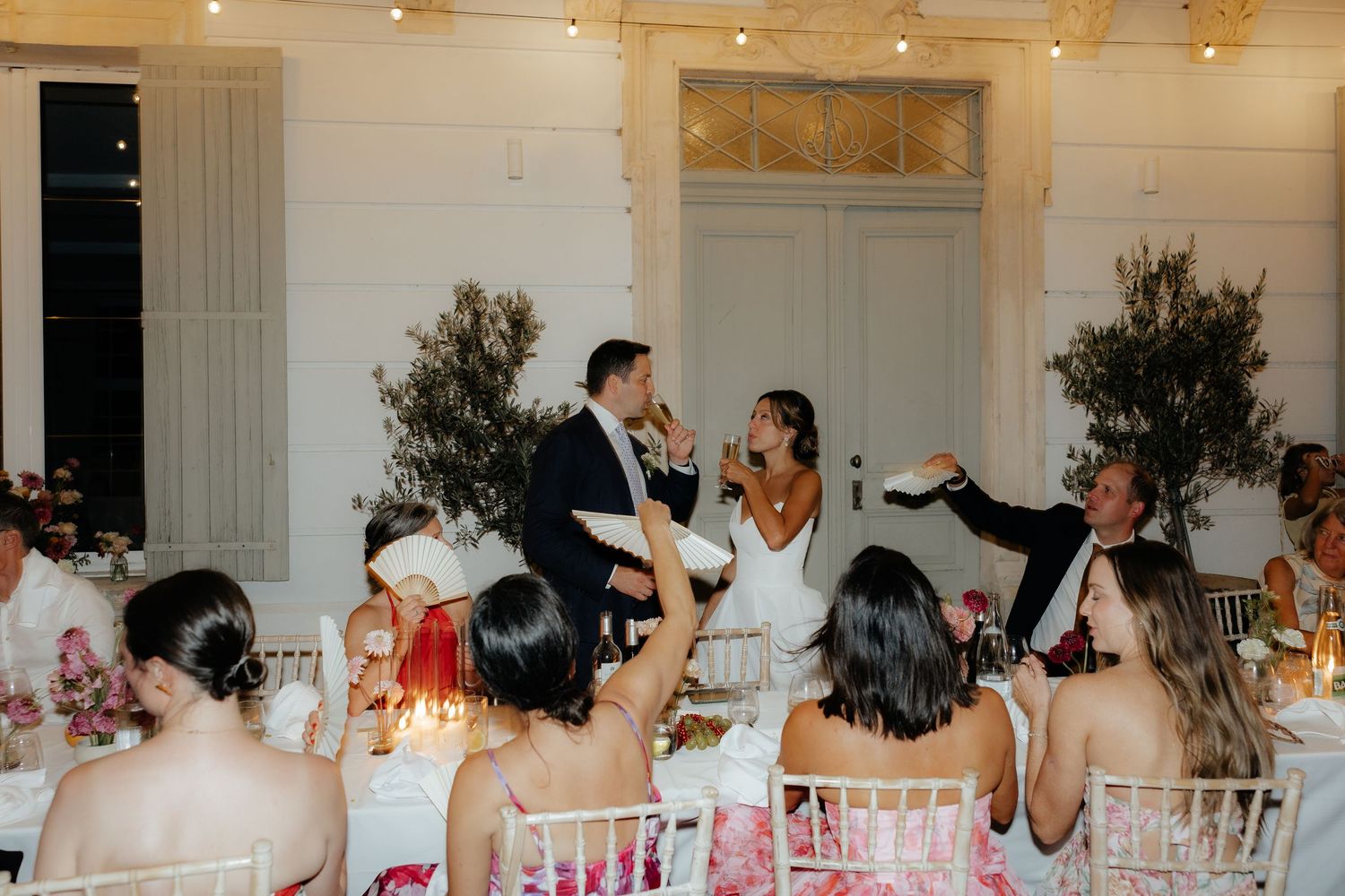 Wedding reception guests raise their glasses in a toast while seated at decorated banquet tables.
