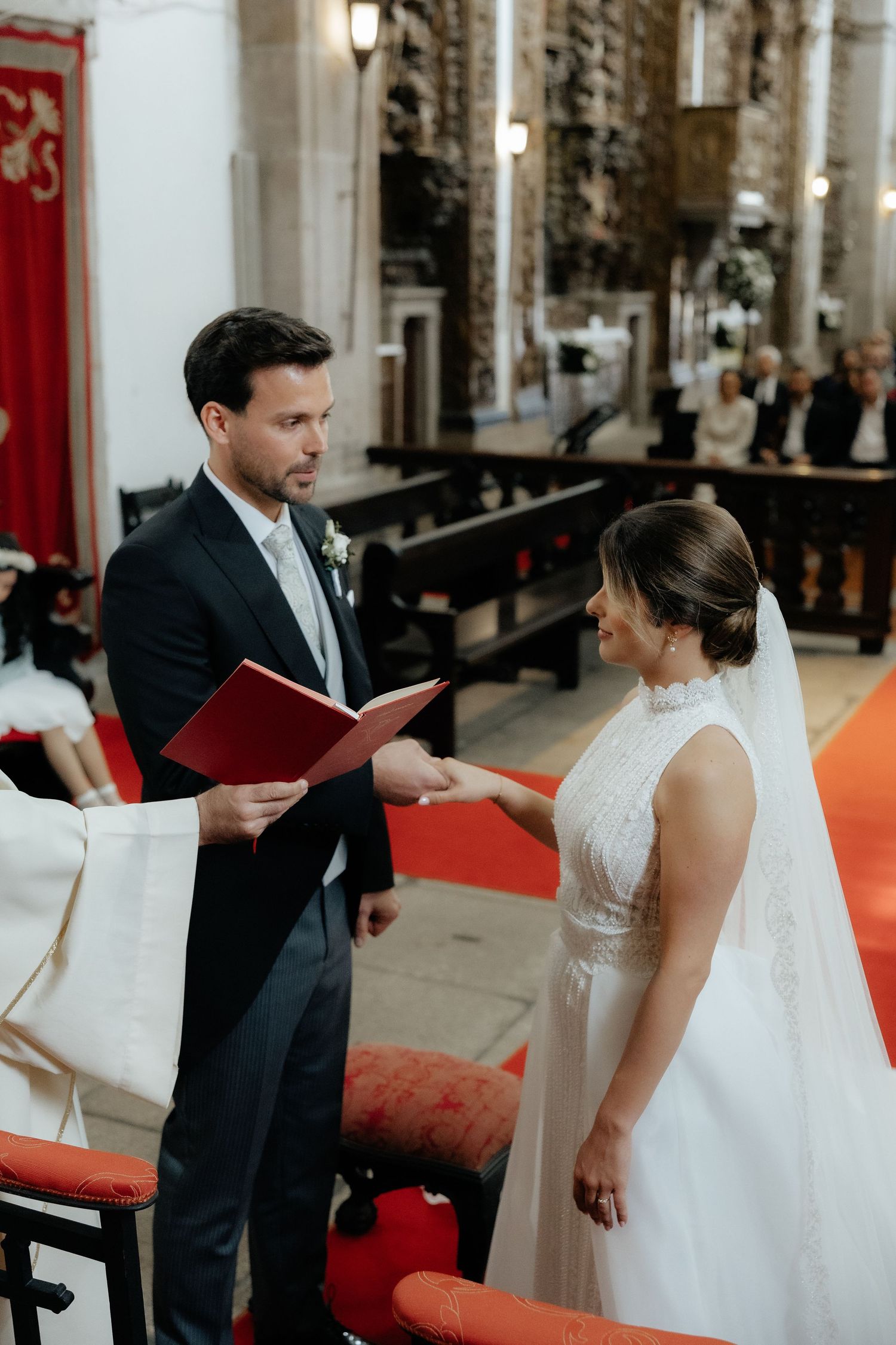 Groom reads from red book while bride smiles during church wedding ceremony exchange.