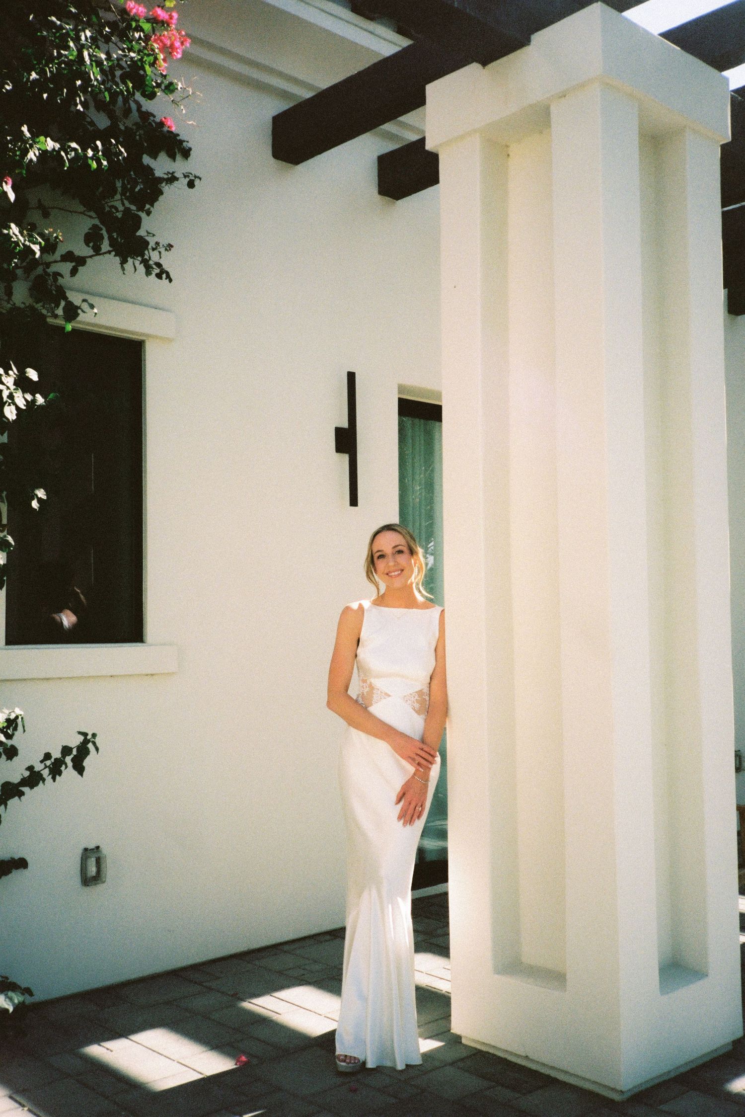 Elegant bride in a sleek white gown poses by a white architectural column in warm sunlight.