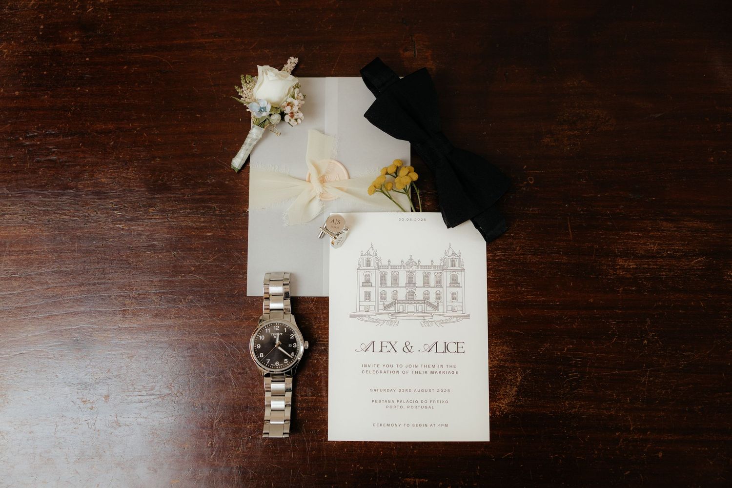 Wedding flat lay featuring an invitation for Alex and Mike, a silver watch, boutonniere, and ribbon on a dark wood surface.
