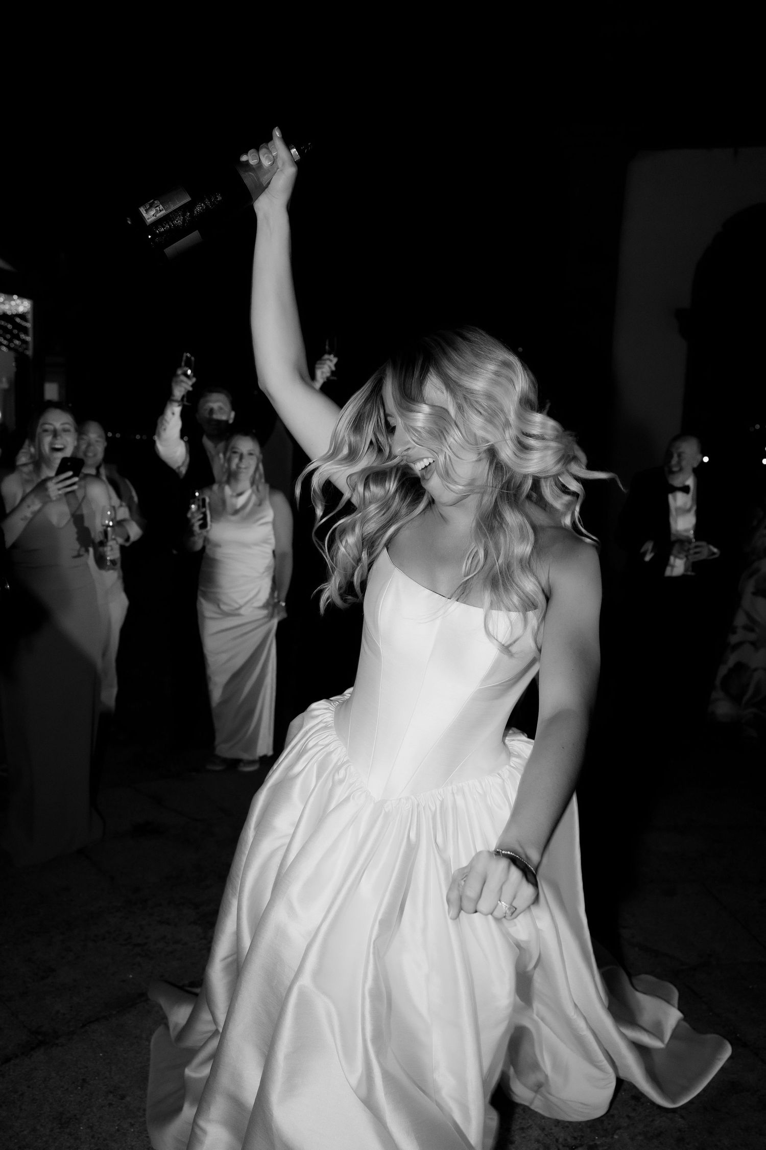 Black and white photo of bride dancing joyfully in wedding gown holding champagne bottle aloft.