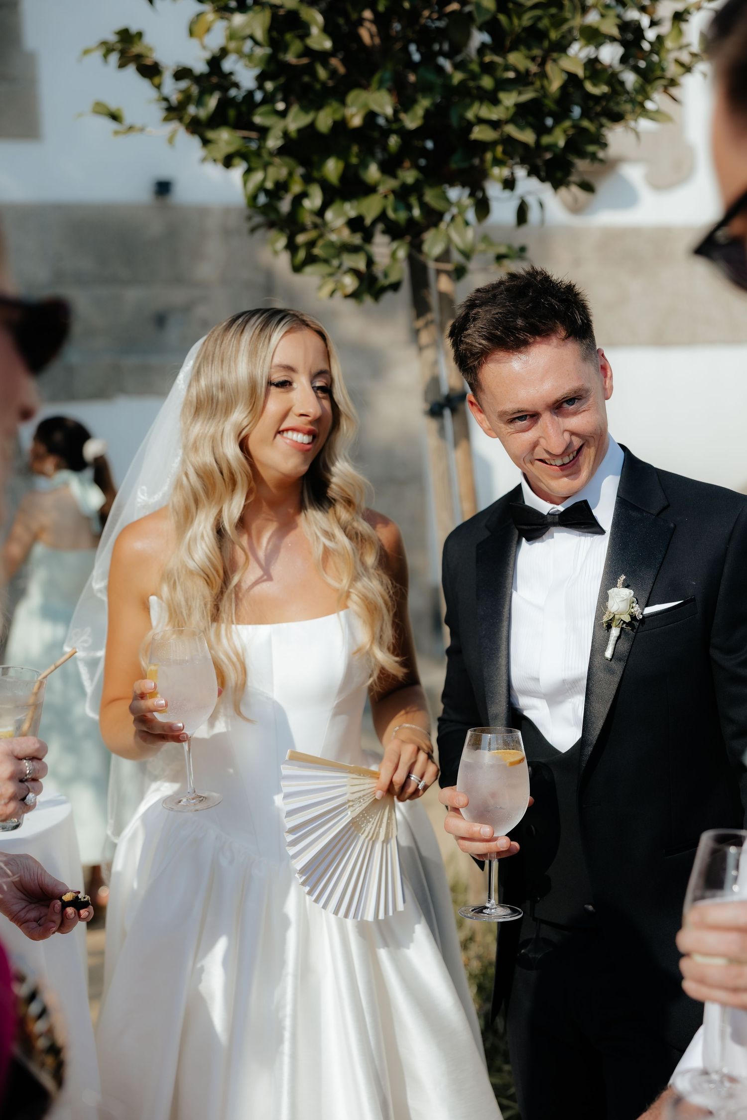 Smiling bride in strapless white gown holding fan and champagne with groom in black tuxedo outdoors.