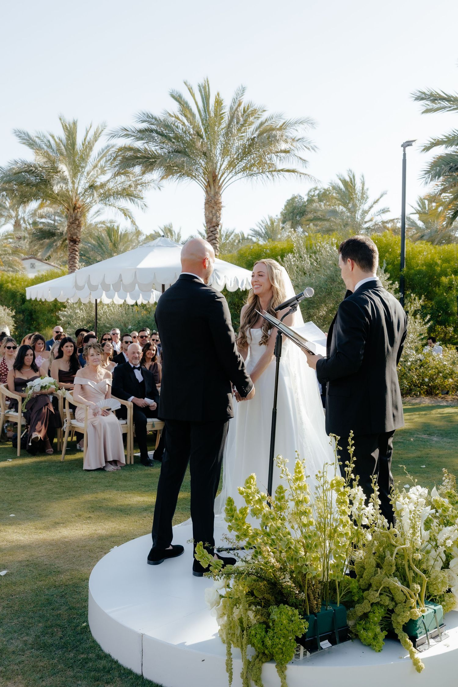 Bride in white gown smiles during outdoor wedding vows on elevated platform with lush greenery and palm trees behind.