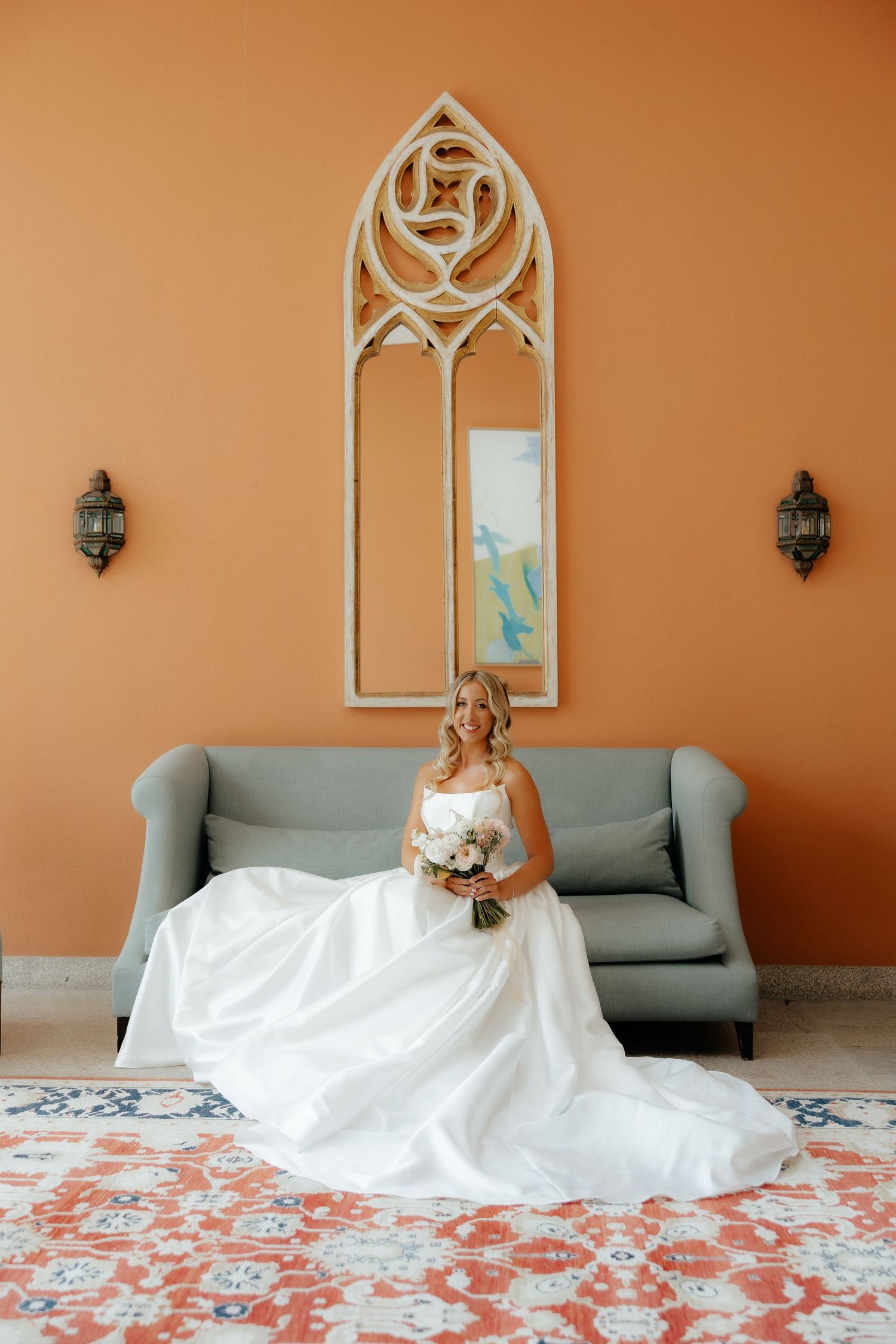 A wedding dress flows gracefully across a patterned floor in front of a gray sofa and arched window decor.