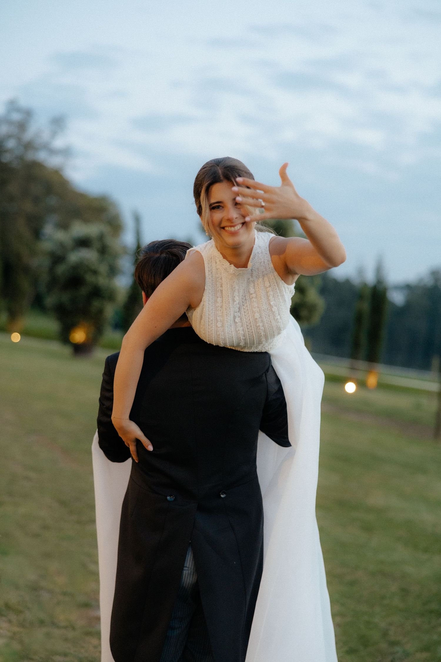 Groom carrying bride on his back outdoors as she laughs and shows her wedding ring.