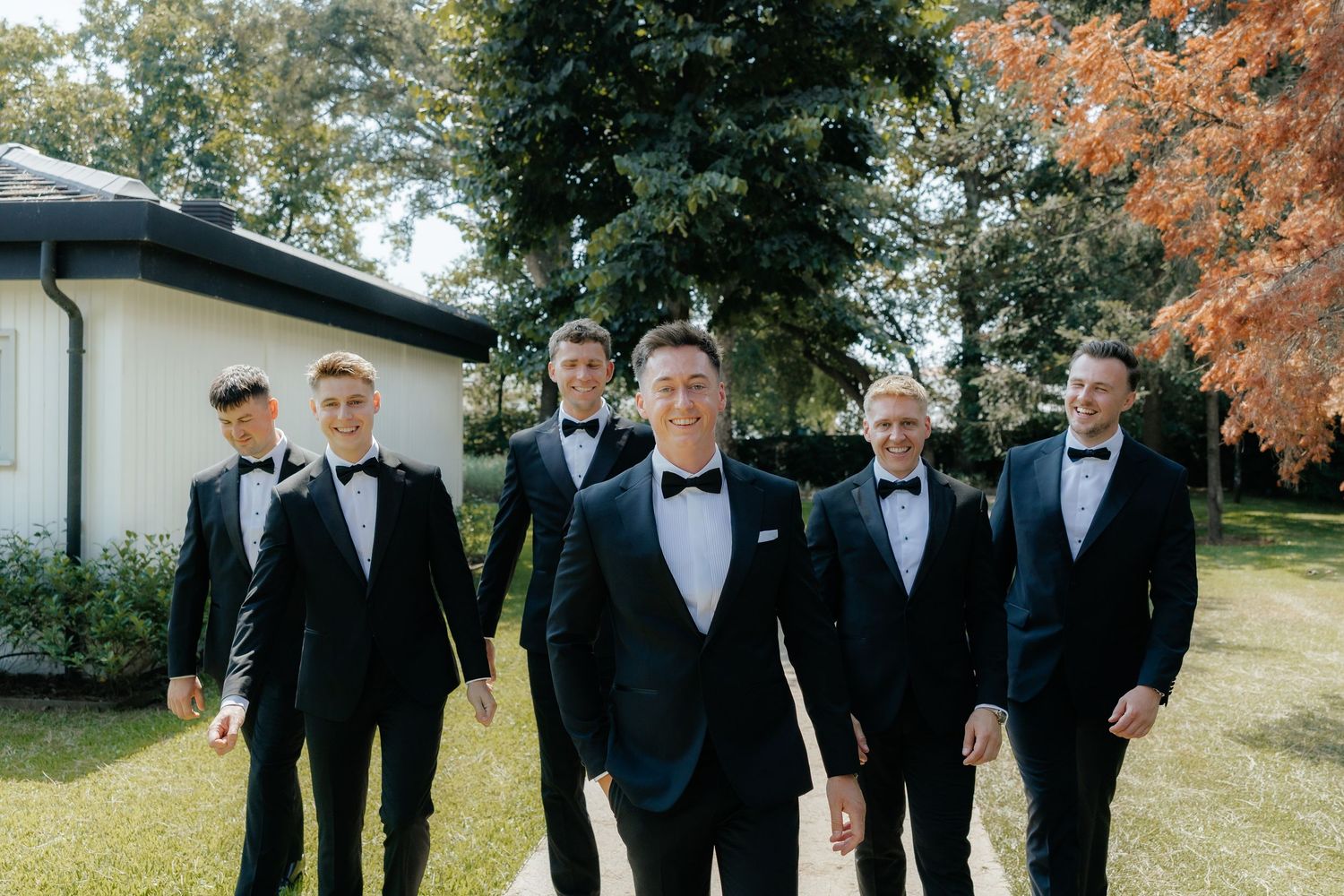 Groom and five groomsmen in matching black tuxedos posing outdoors on a sunny day with green trees behind them.