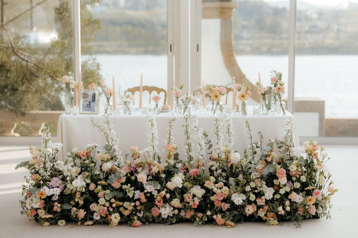 Elegant wedding head table adorned with lush pink and white floral arrangements and tall candles by a window.