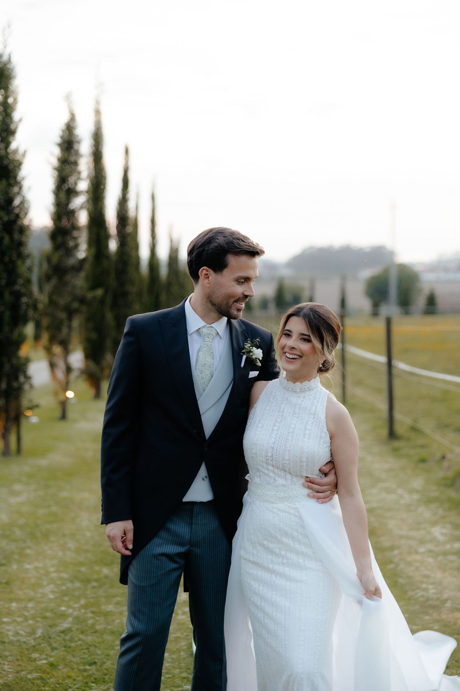Smiling bride in high-neck beaded gown and groom in navy suit walking together along cypress tree-lined path.