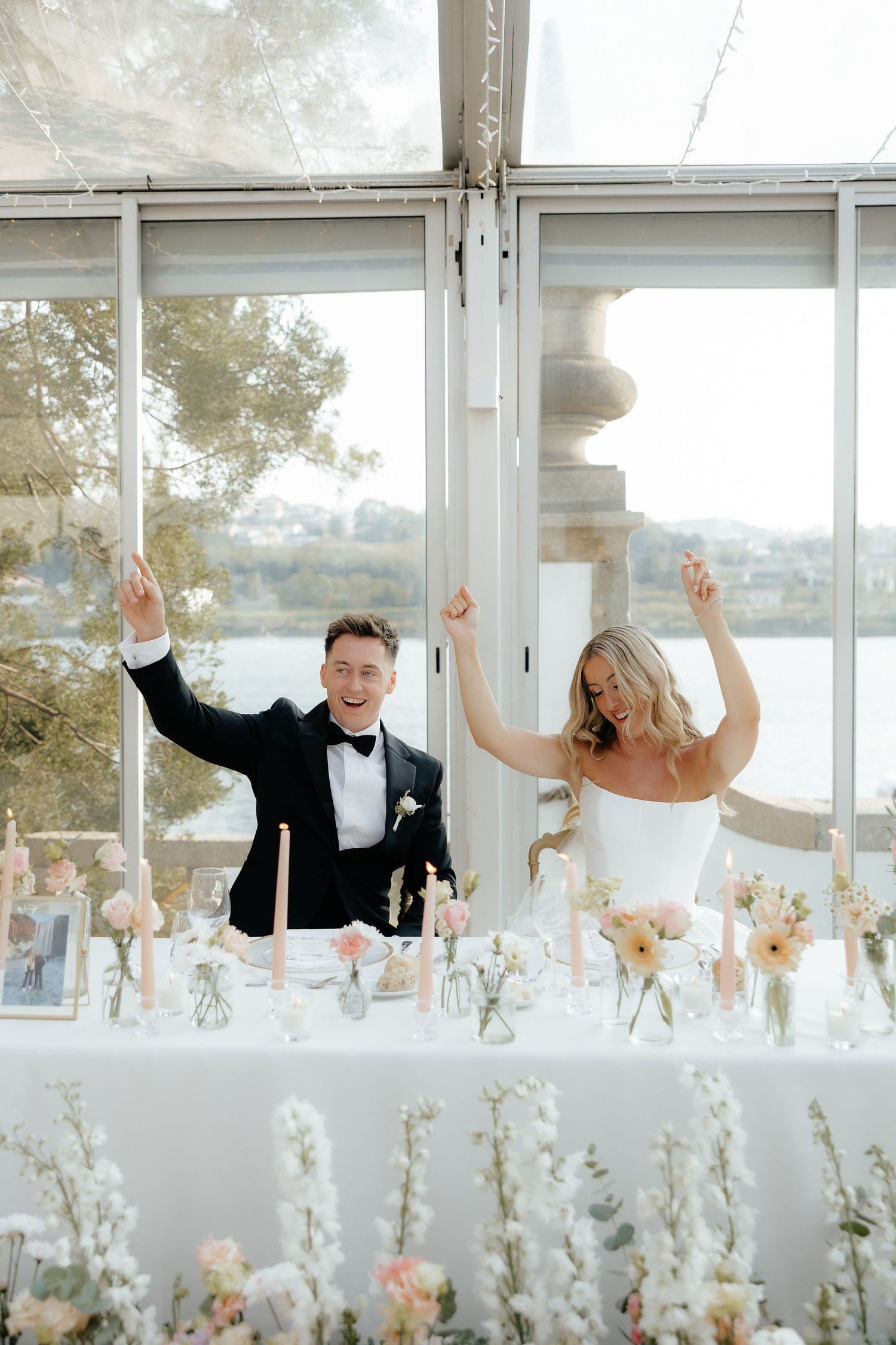 Wedding couple peeks playfully above table decorated with white floral centerpieces in bright reception venue.