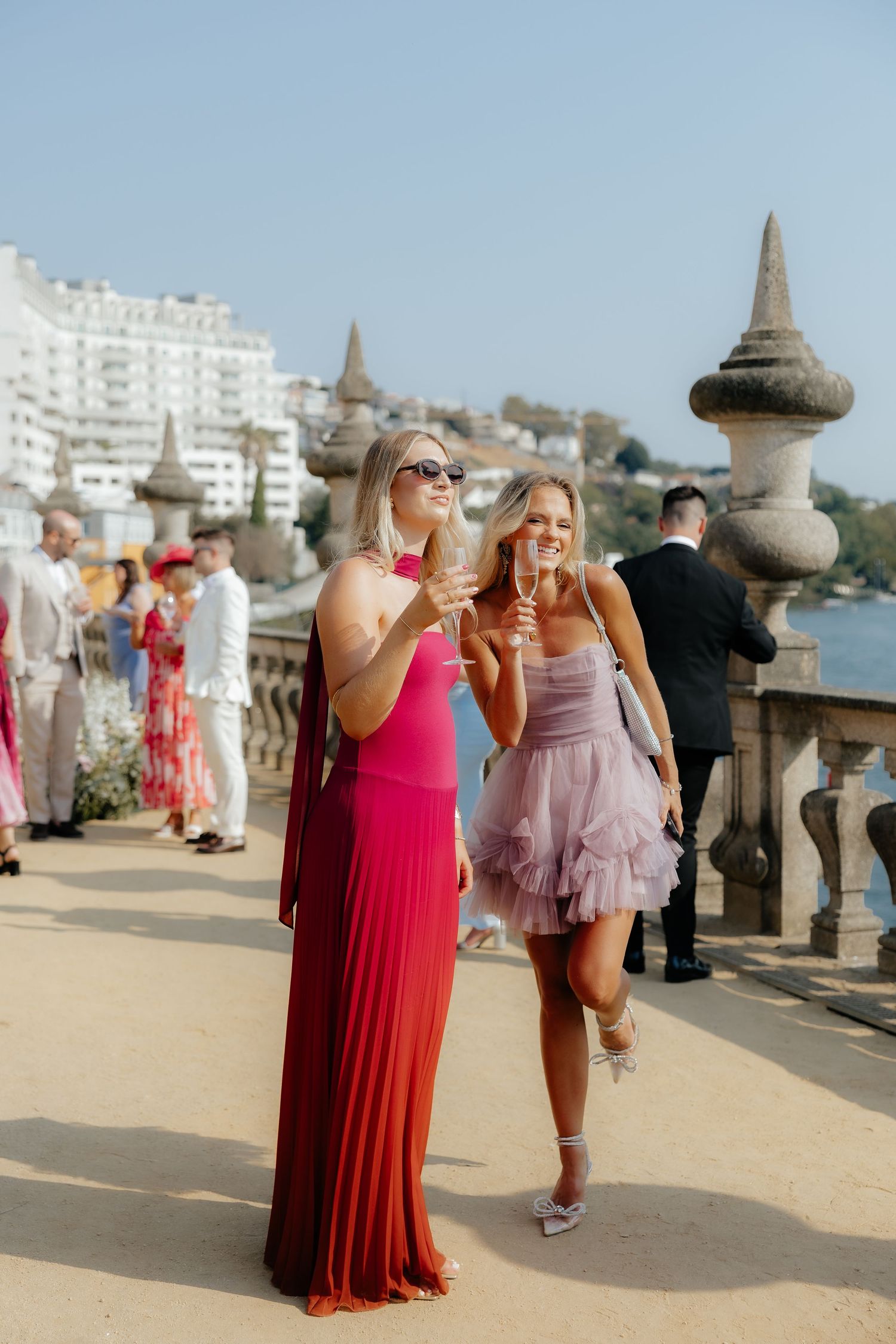 People in formal attire gather on a seaside promenade with decorative stone pillars and city buildings in background.