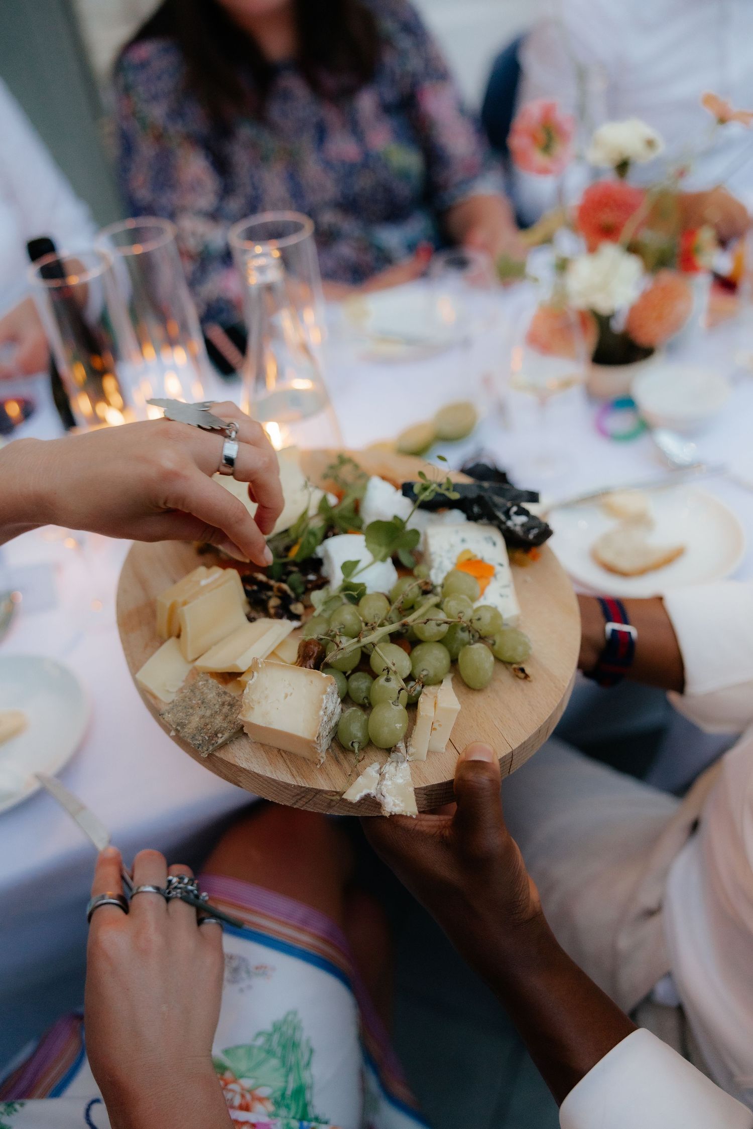 Hands reaching for food on a wooden cheese board with grapes and other appetizers at a candlelit gathering.