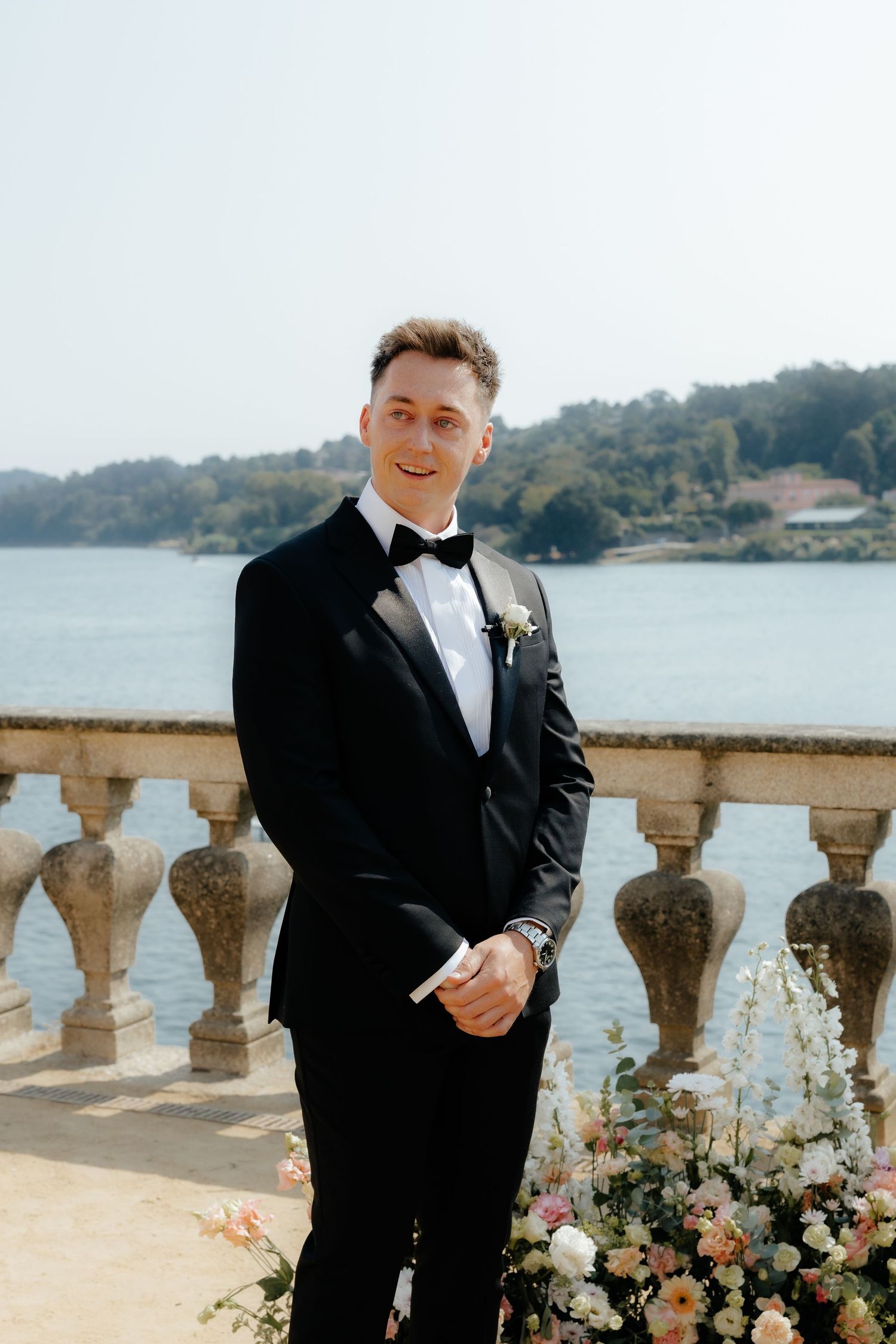 A groom in a black tuxedo stands by stone balustrades overlooking a waterfront view during a wedding ceremony.