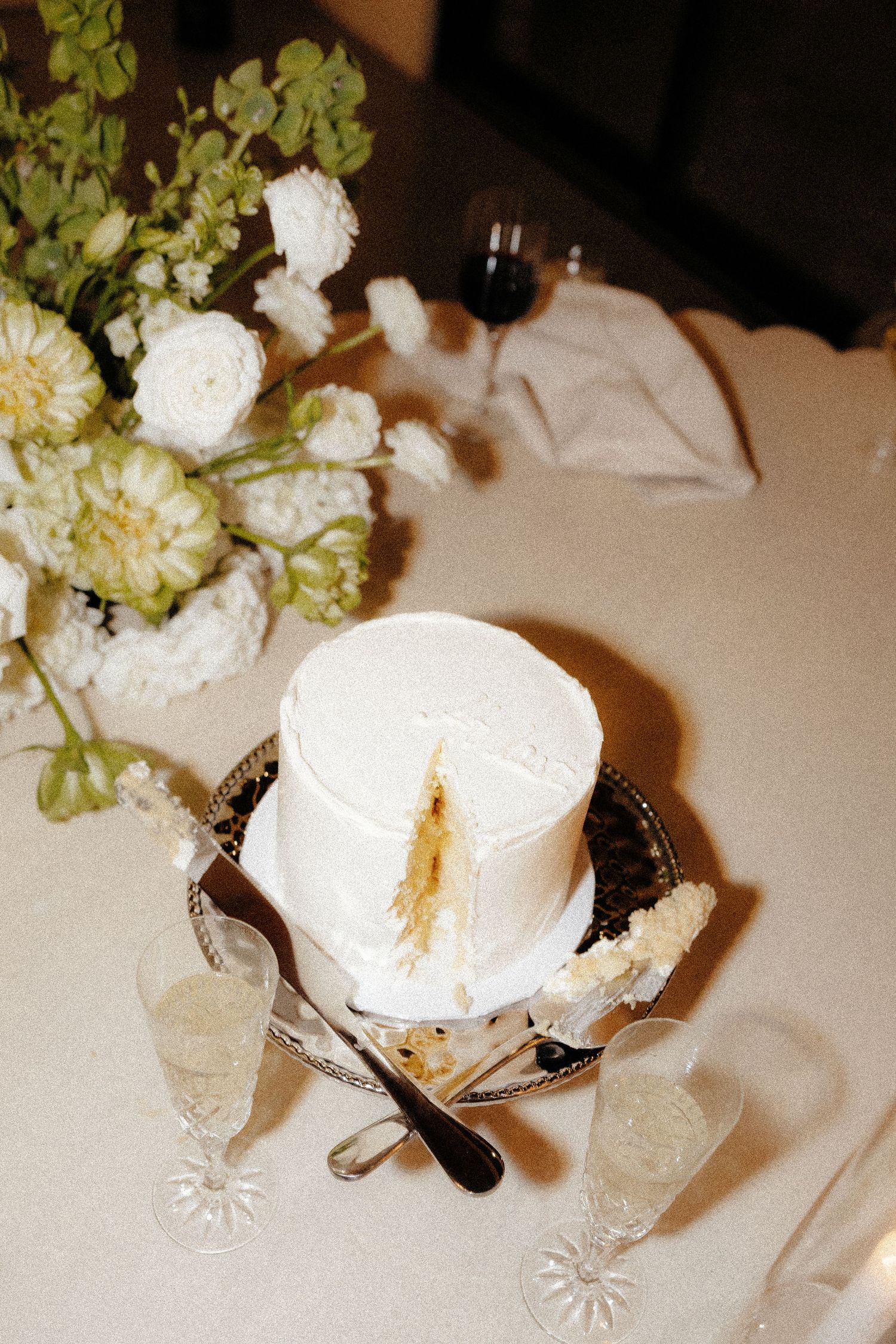 Elegant single-tier white wedding cake with a dried pampas grass topper displayed beside white floral centerpiece on reception table.