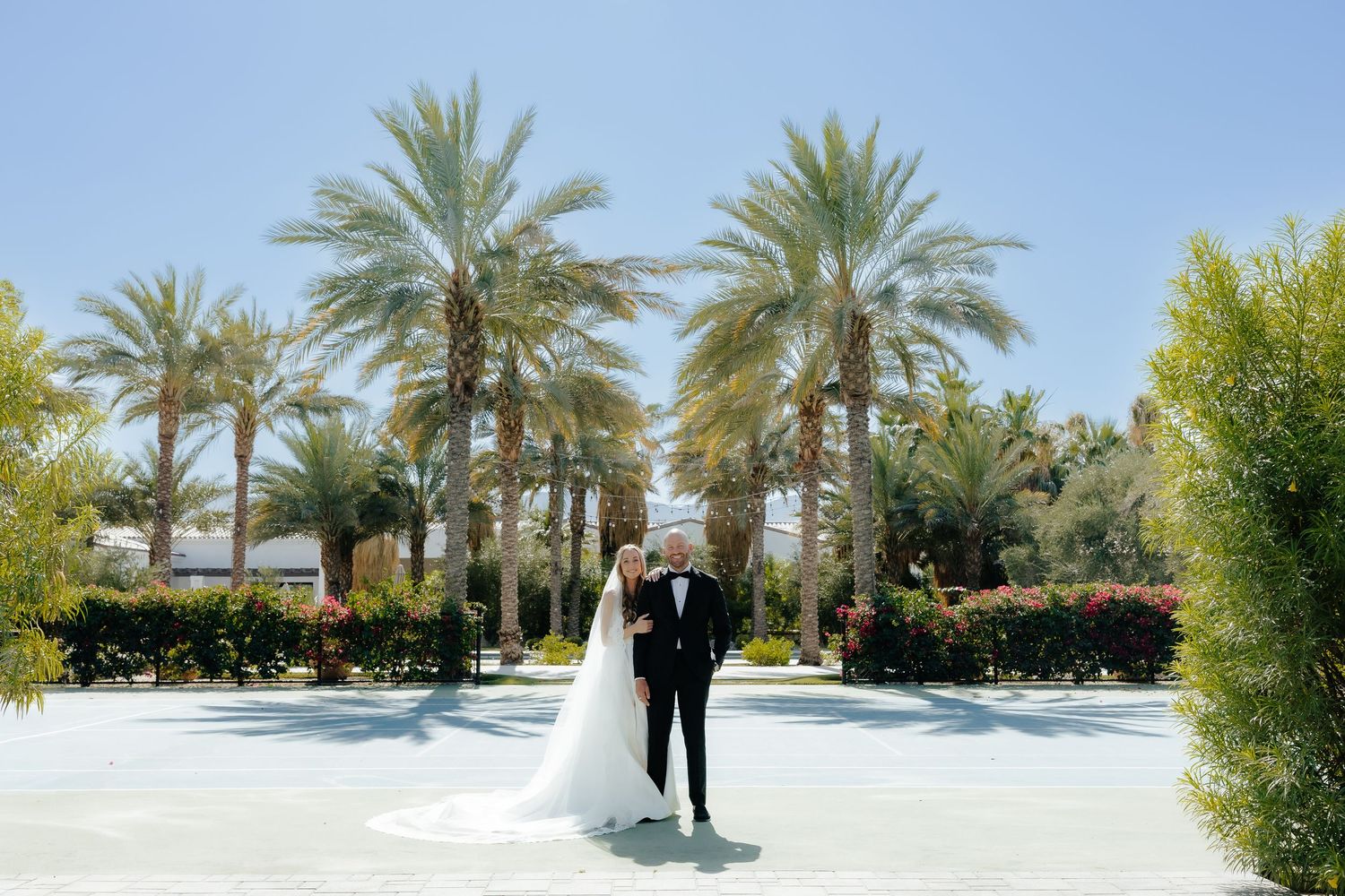 Bride and groom standing together in an open plaza surrounded by tall palm trees.
