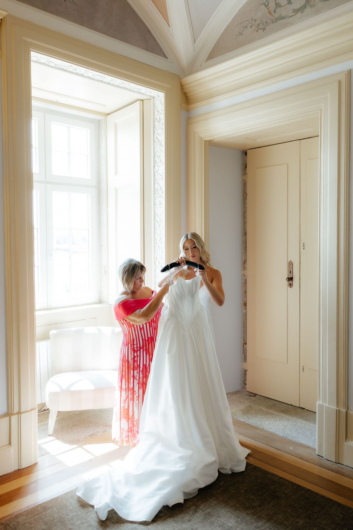 A white wedding dress being fitted in a sunlit room with ornate architectural details and tall windows.