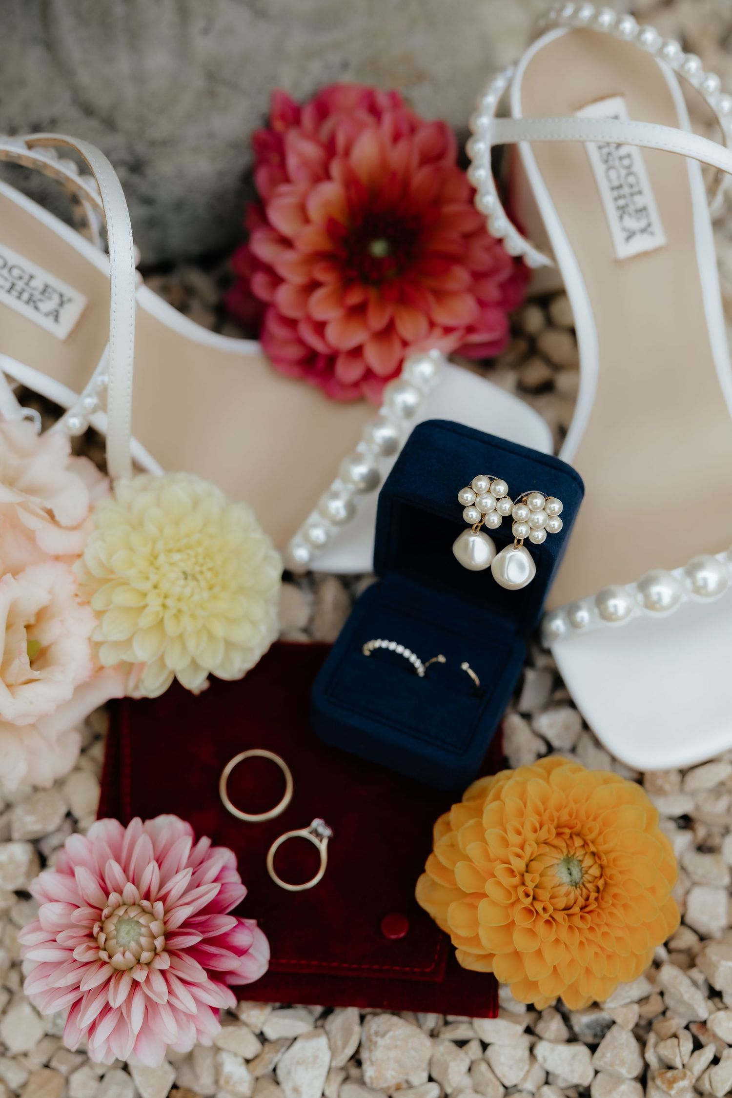 Wedding rings displayed with colorful dahlias and pearl accessories on a textured surface.