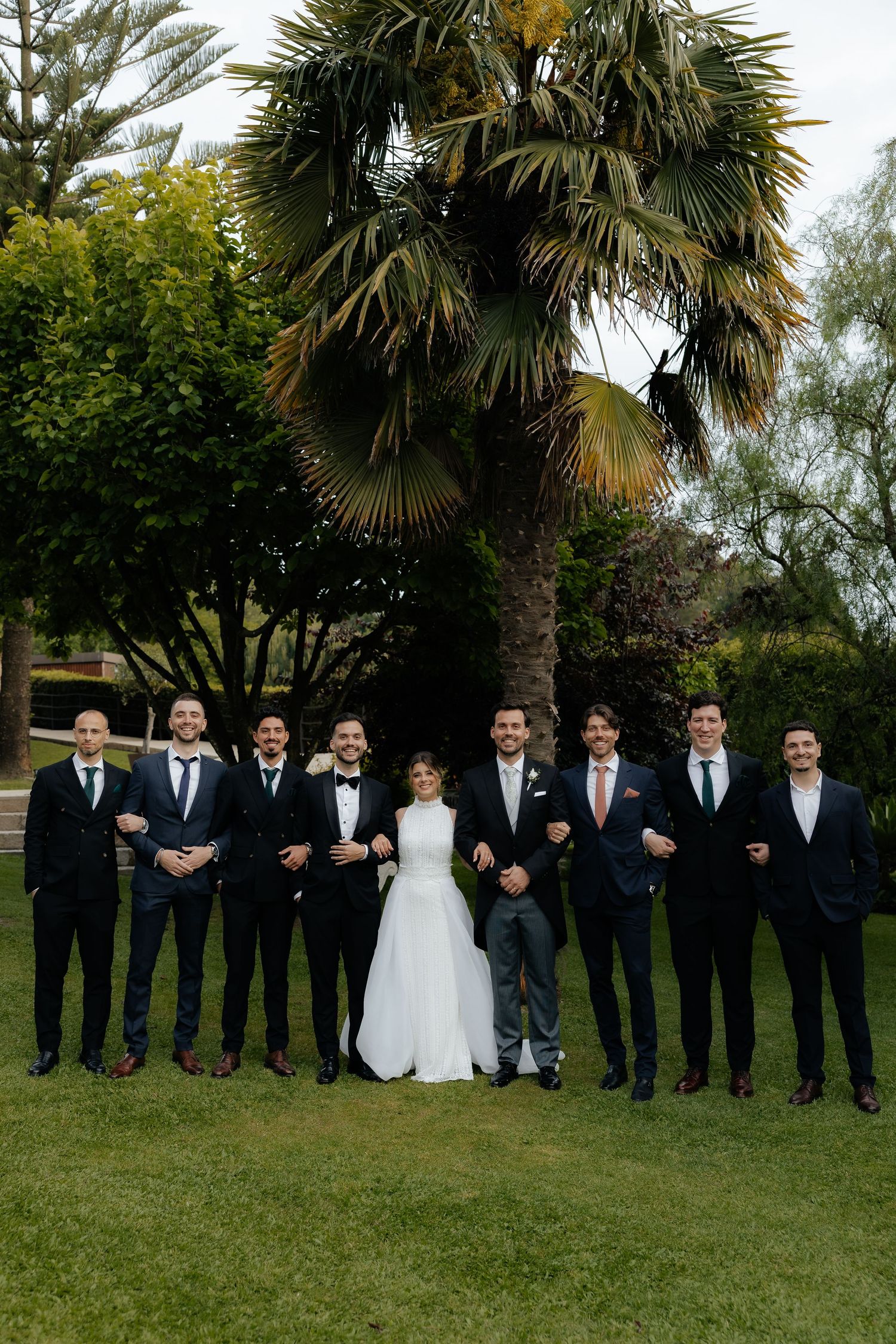 Bride poses with groomsmen in dark suits on green lawn with tropical palm trees behind them.