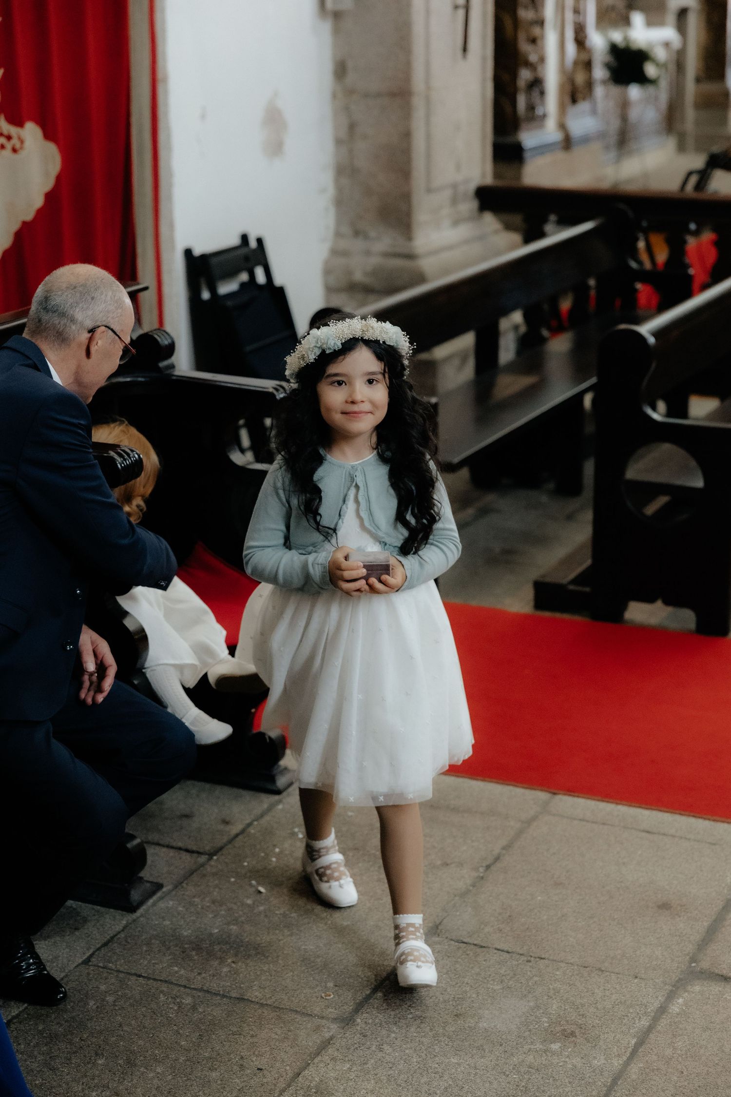 Young flower girl in white tulle dress and floral crown stands on red carpet.