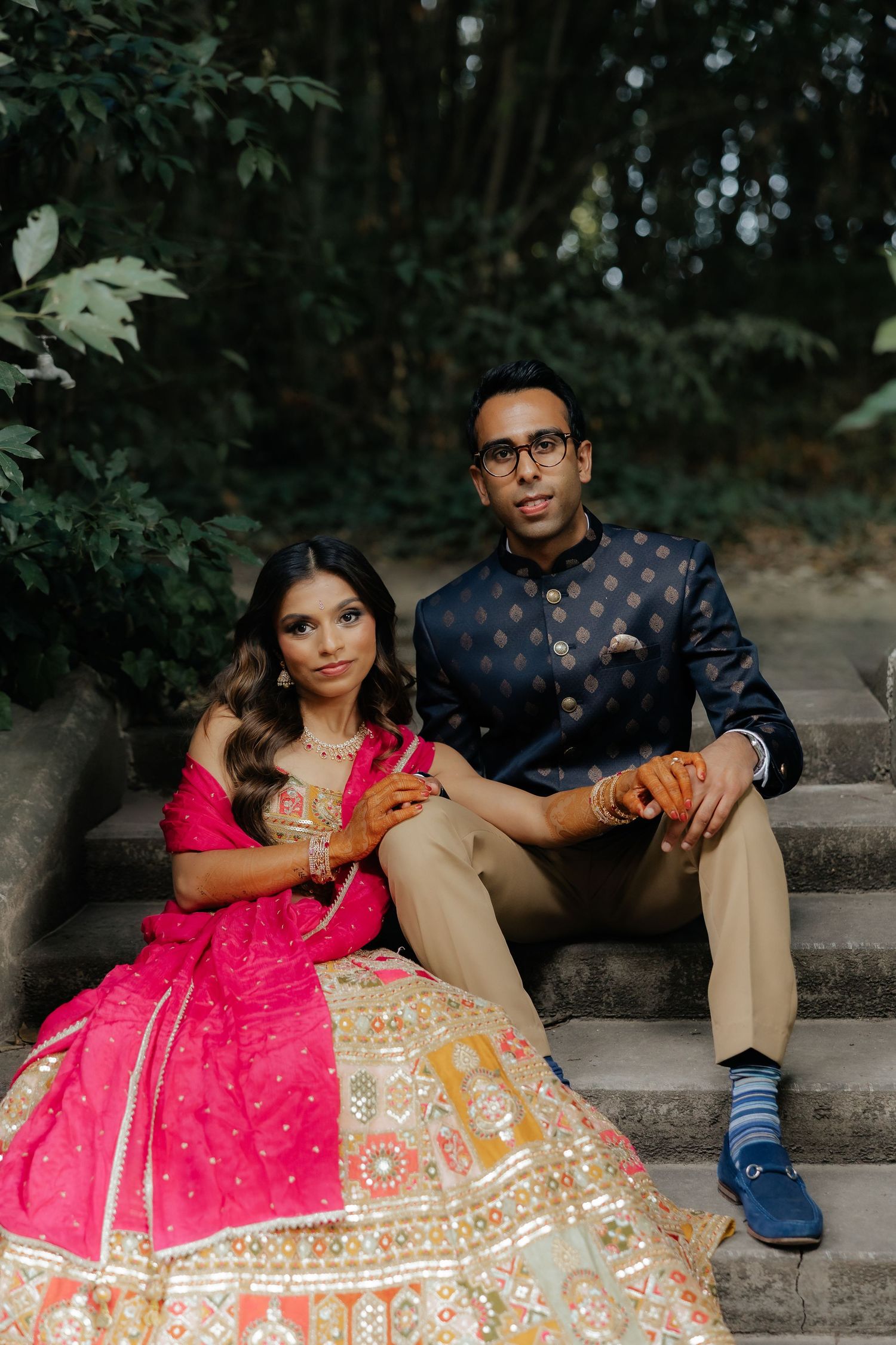 Couple in traditional Indian wedding attire sitting on stone steps surrounded by lush greenery.