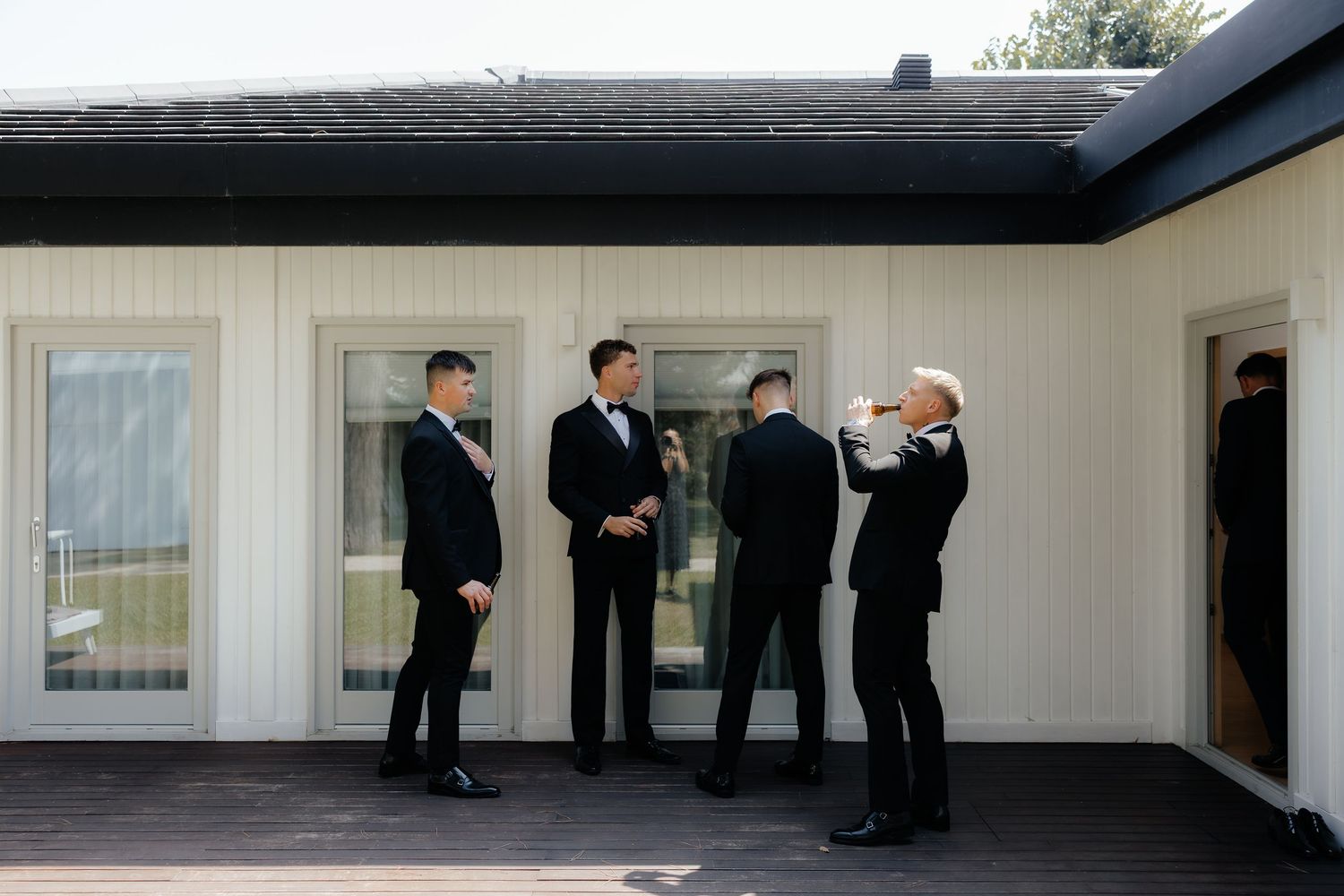 Groomsmen in black tuxedos getting ready on an outdoor deck of a white building on a wedding day.