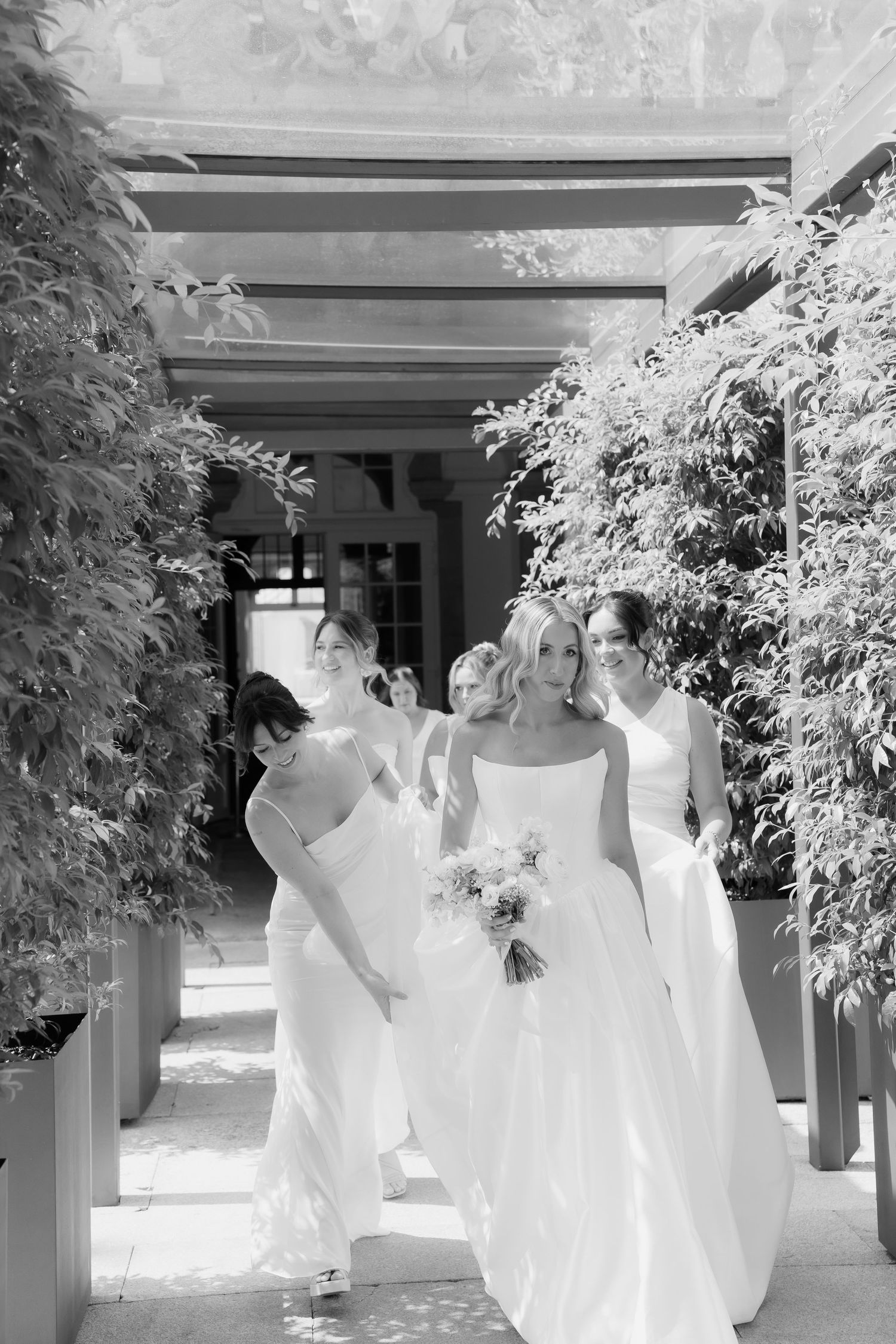 Black and white photo of bride and bridesmaids walking through vine-covered outdoor corridor.