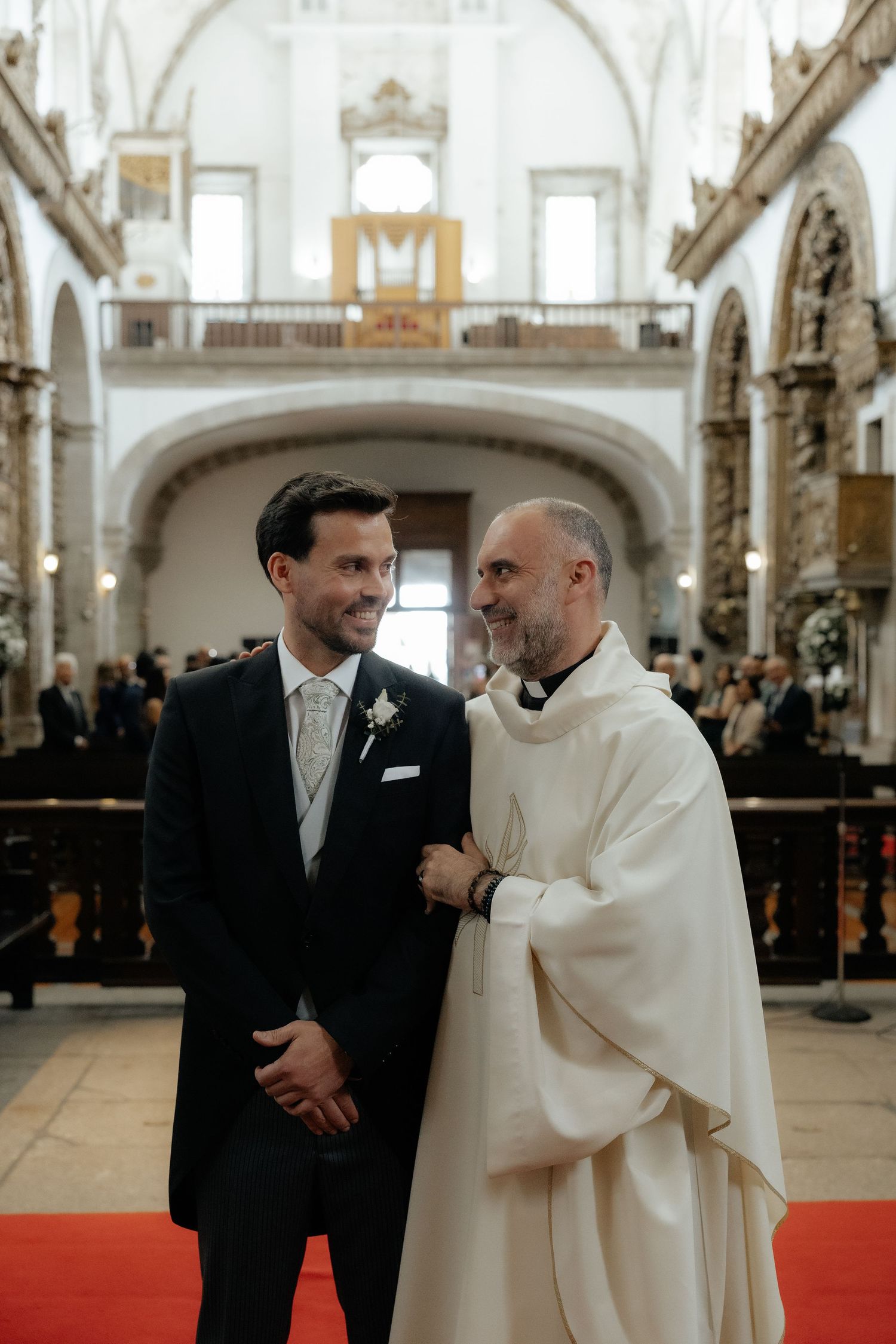 Groom in black suit smiles with priest in white robe inside ornate church.
