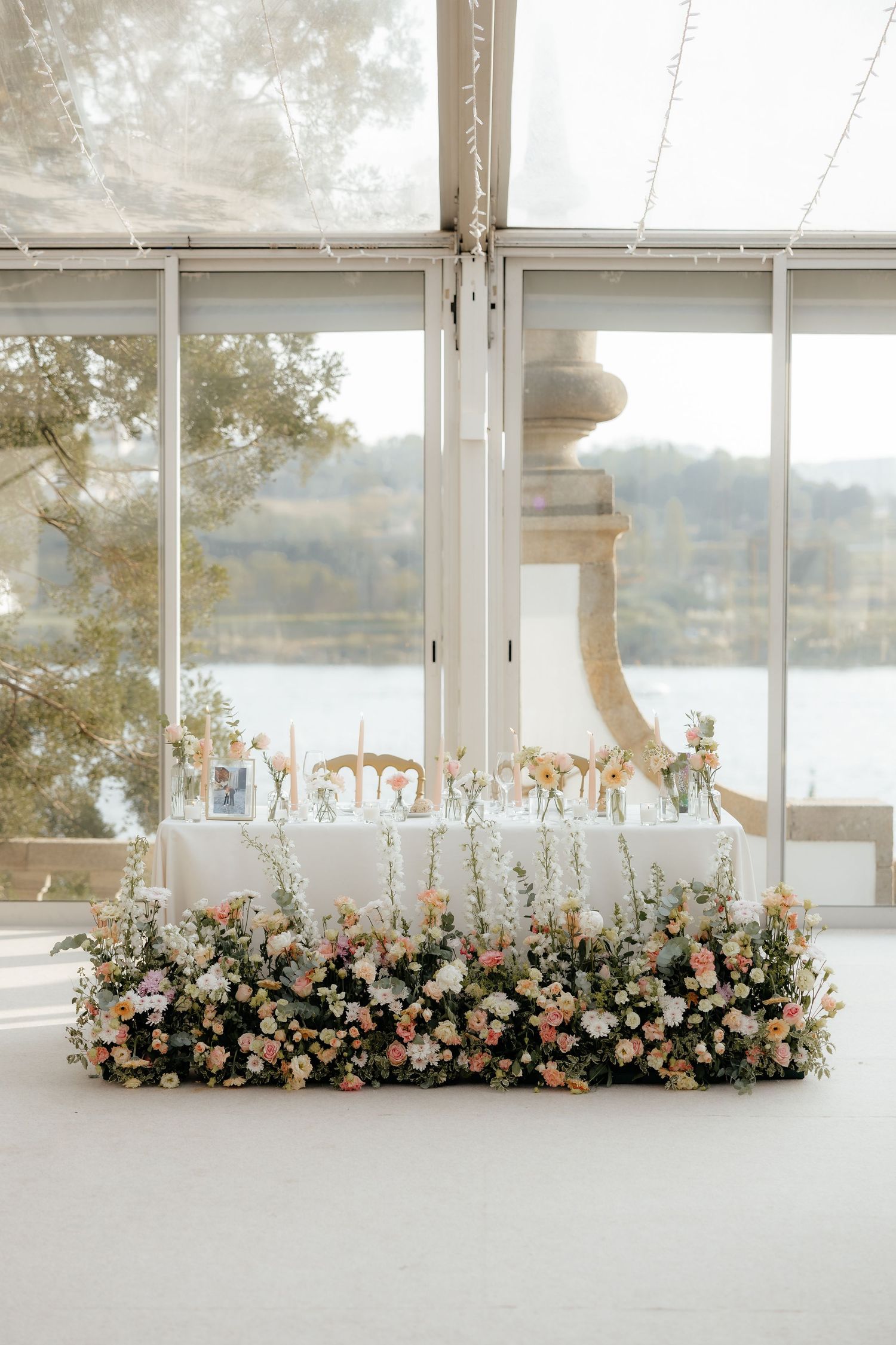 Elegant floral arrangements with peach and white flowers line a bright window overlooking a serene waterfront view.