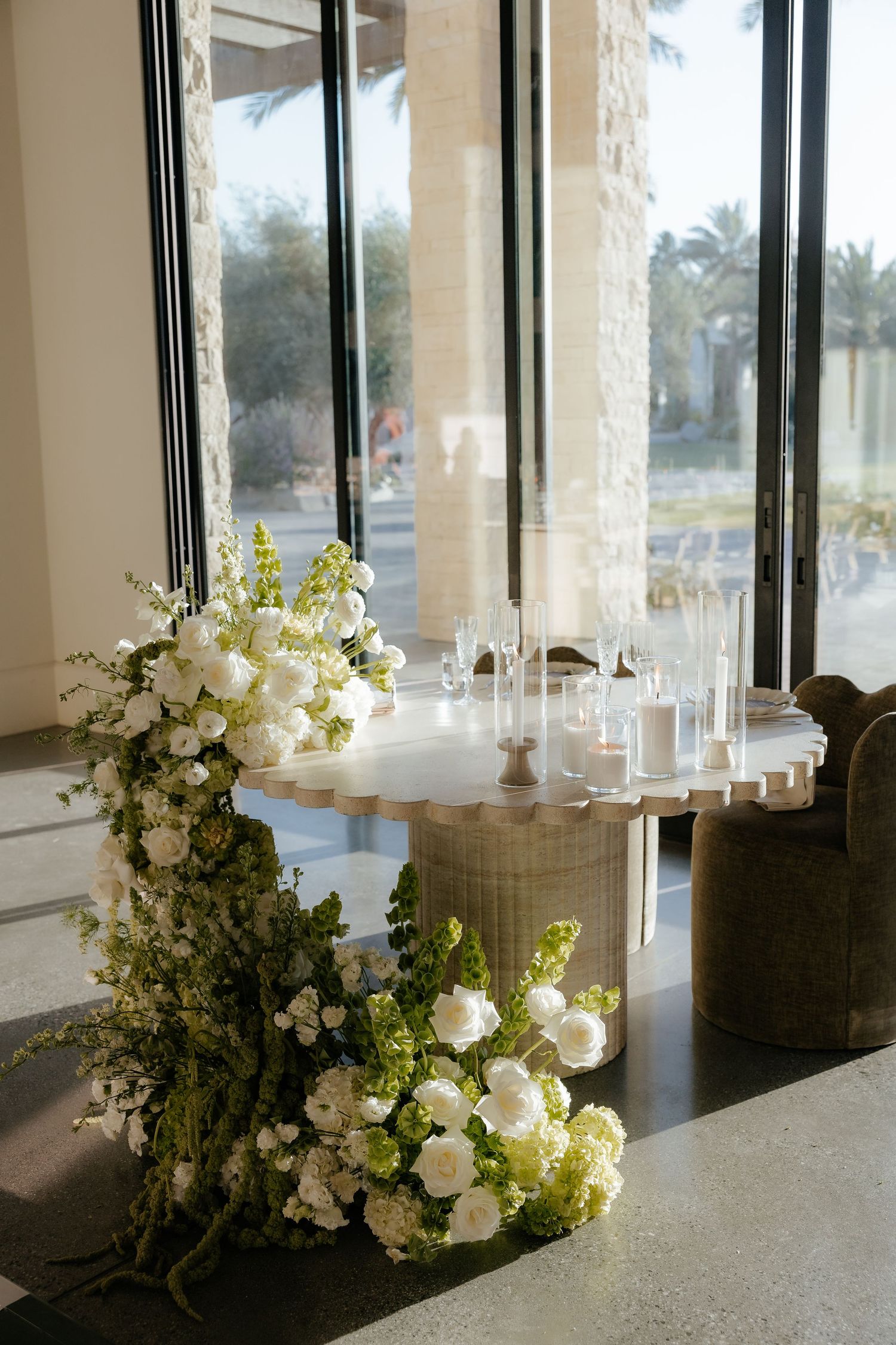 Sweetheart table adorned with cascading white and green floral arrangements beside floor-to-ceiling windows.